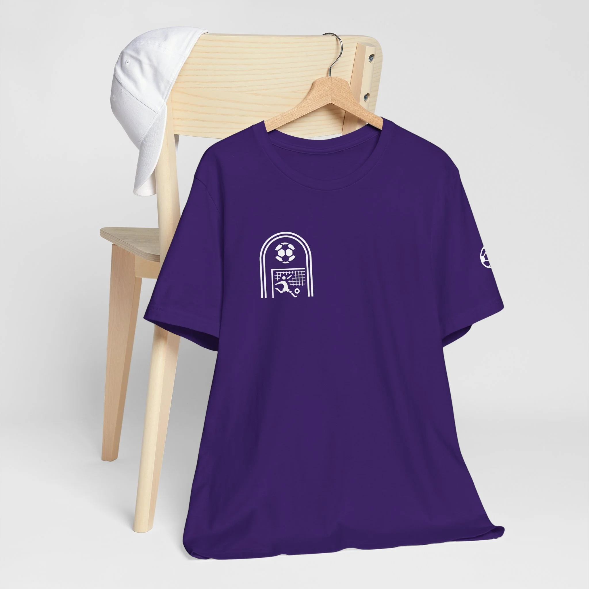 Purple T-shirt with soccer-themed graphics hanging on a wooden hanger, with a white cap draped over the back of a wooden chair in the background.