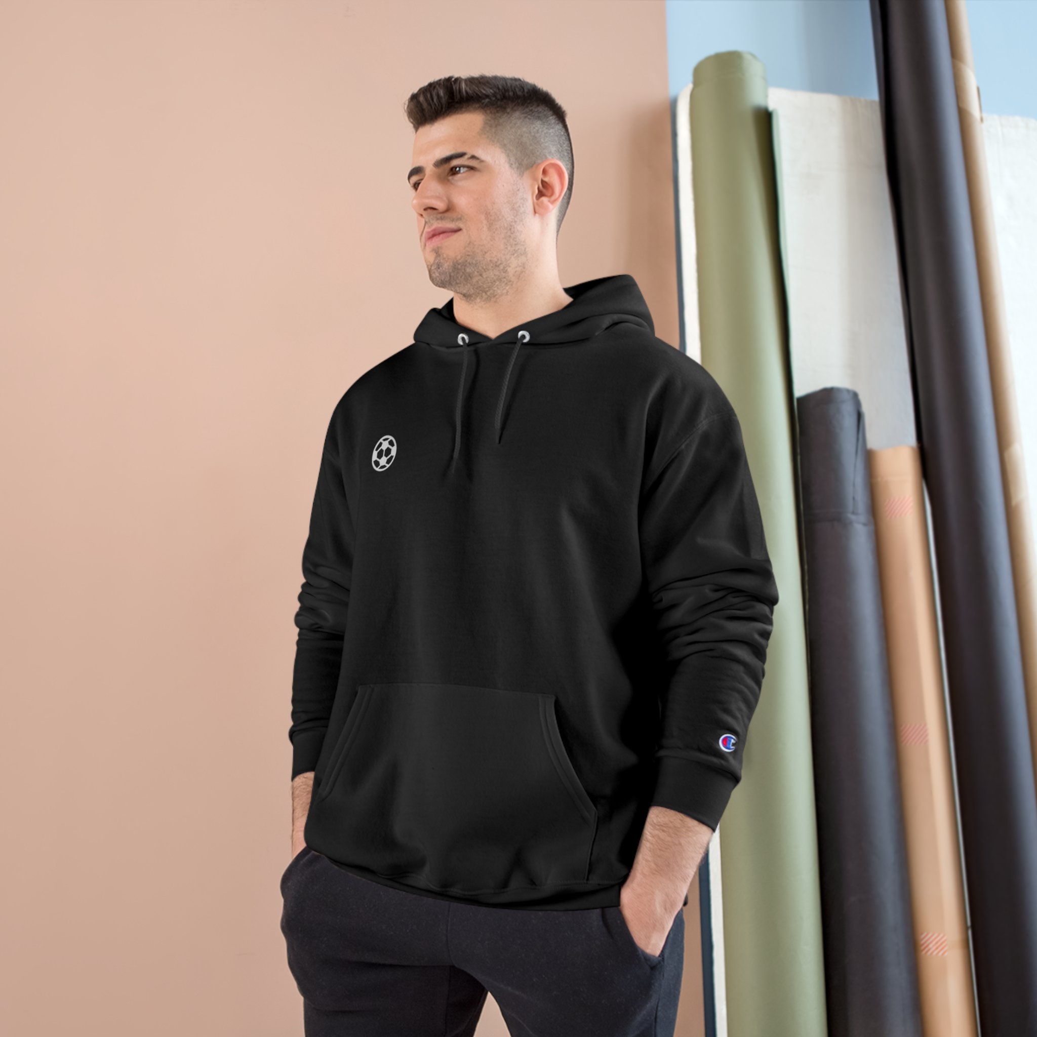 Young man wearing a black hoodie with a sports logo on the chest and sleeve, standing indoors with rolls of wrapping paper or wallpaper behind him.