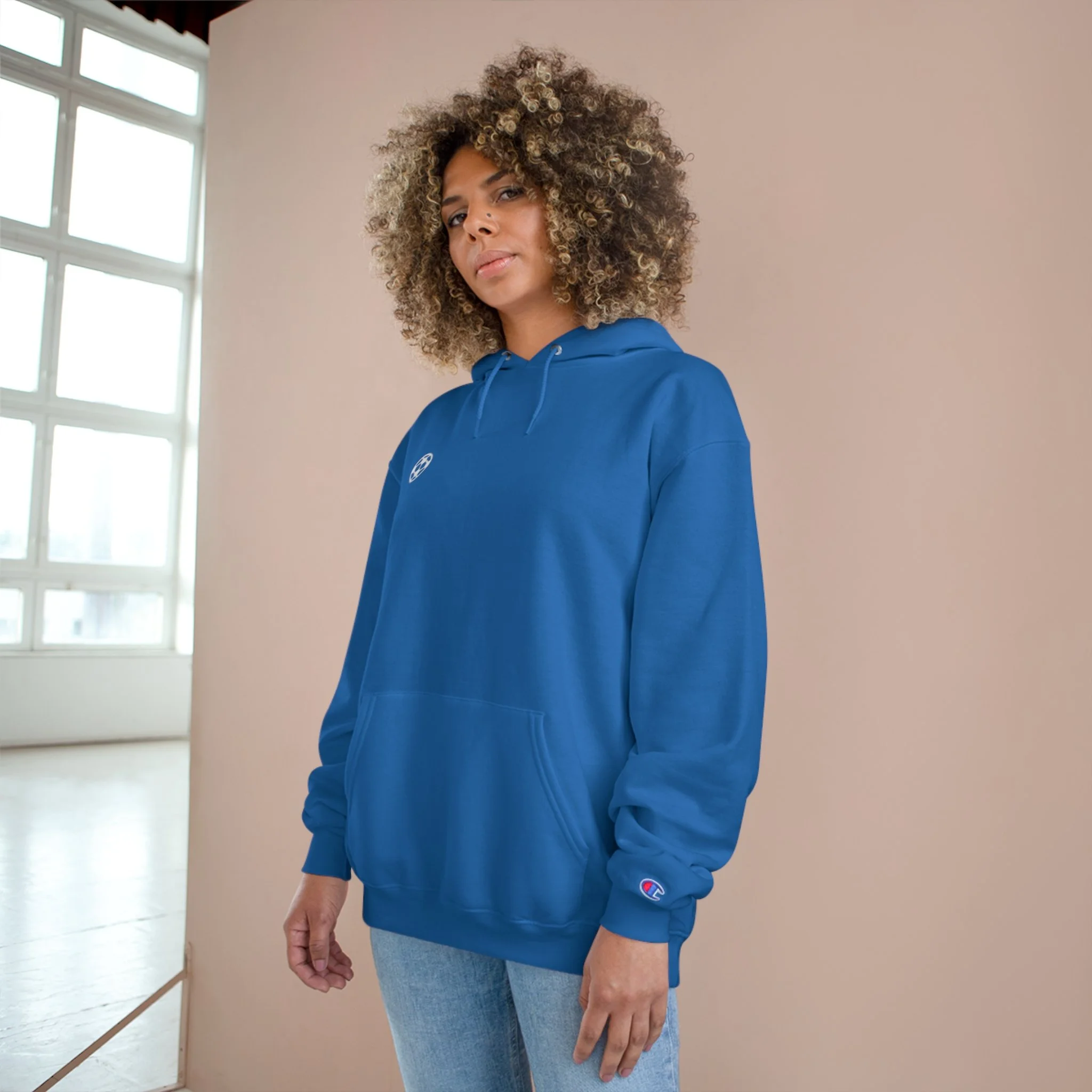 A woman with curly hair wearing a blue hoodie and light jeans, standing indoors by a pink wall and a large window in the background.