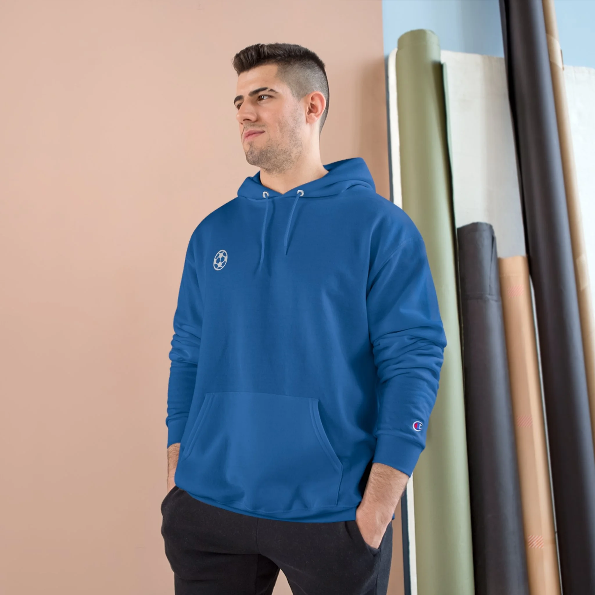 A young man with dark hair and a beard, wearing a blue hoodie with a small logo on the chest, standing with hands in pockets against a neutral-colored wall with rolls of fabric in the background.