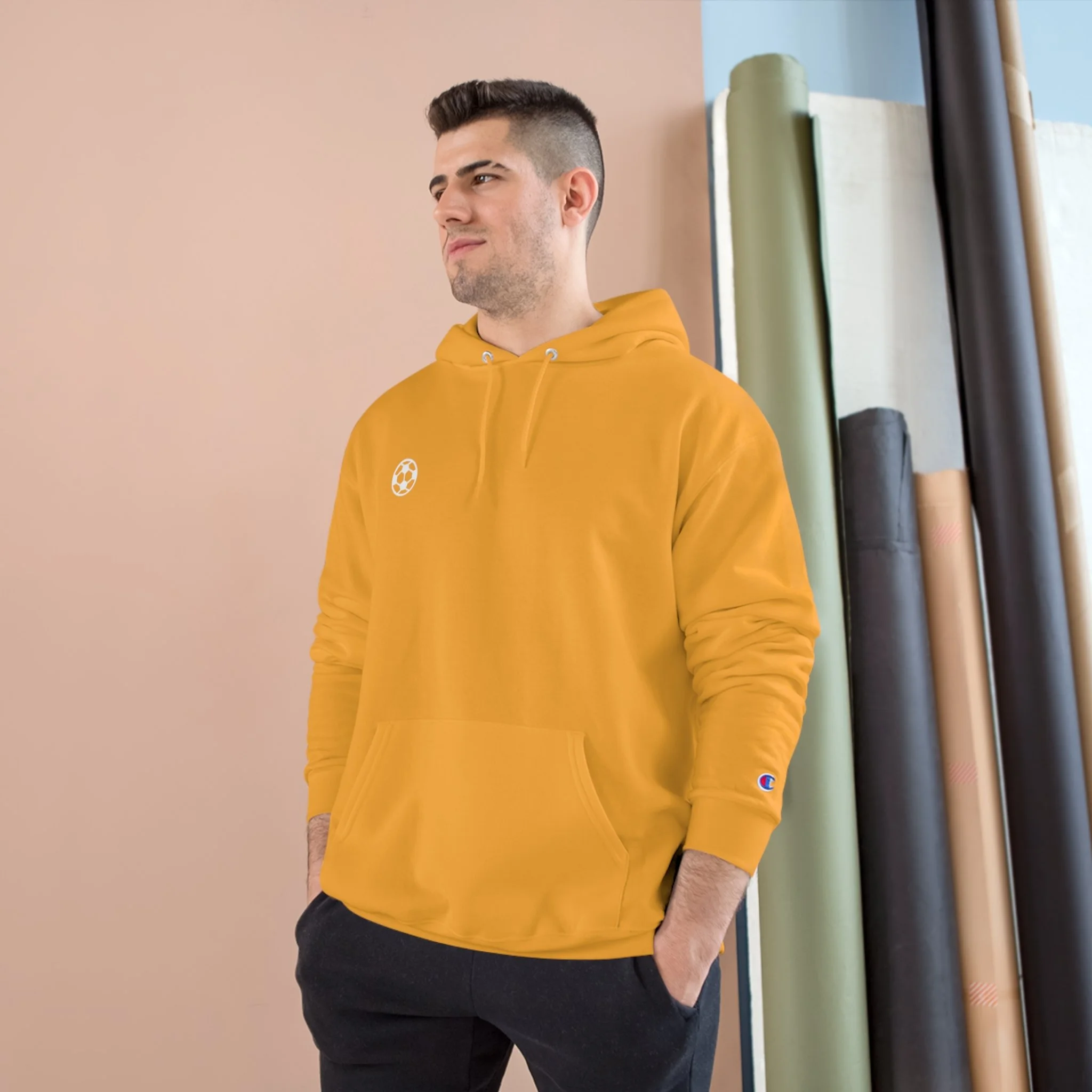 A young man in a yellow hoodie with a small soccer ball logo, standing indoors with hand in pocket, against a background of rolled-up mats.