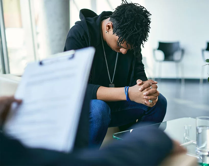 A young man with dreadlocks and a black hoodie sitting with clasped hands during a serious moment in a modern room.