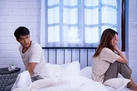 A young man and woman sitting on a bed in a bedroom, facing away from each other, appearing upset or deep in thought.