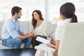 A couple sitting on a couch holding hands during a therapy session with a therapist taking notes.