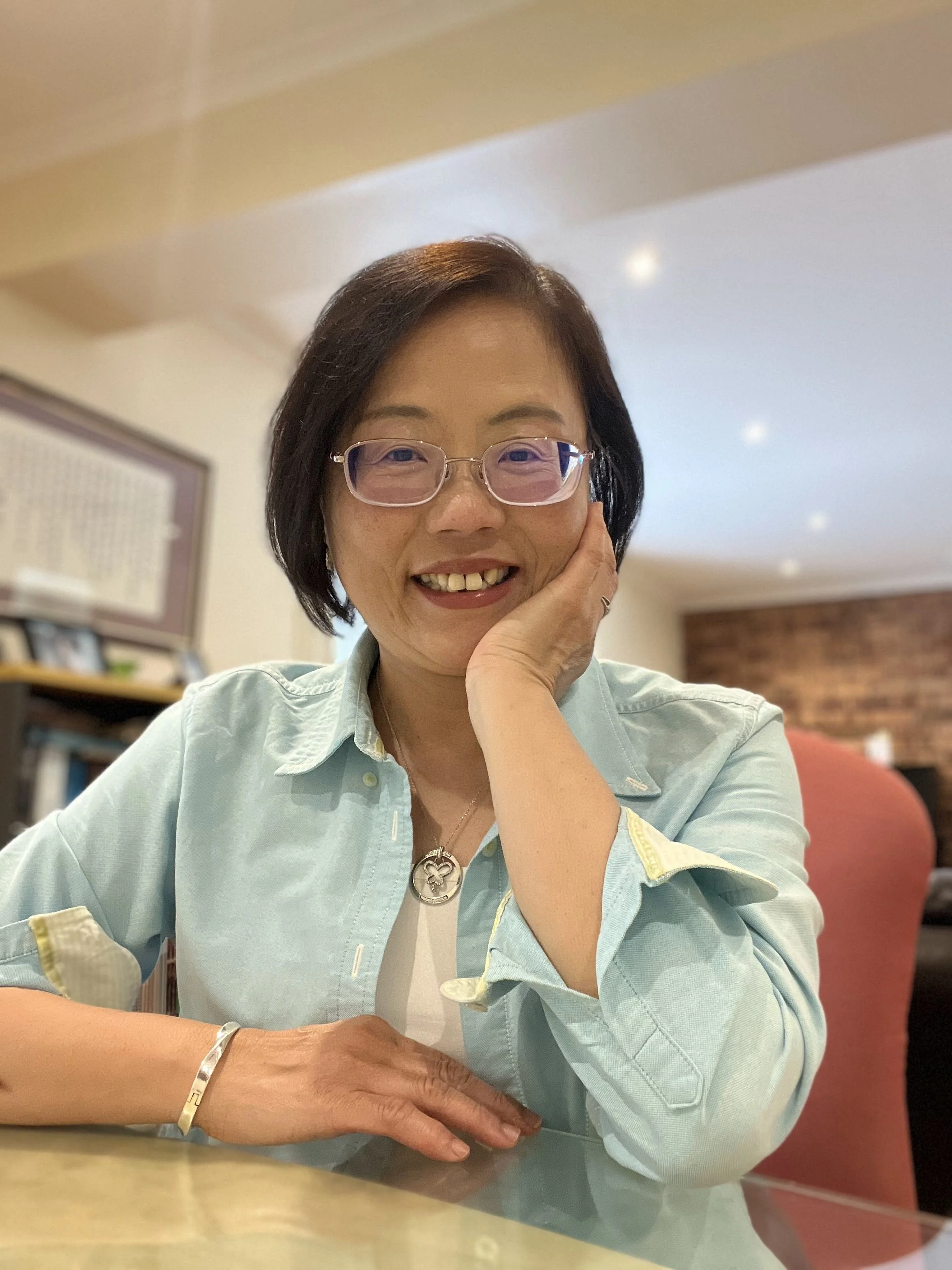 A woman with glasses smiling and resting her face on her right hand, sitting at a table in a cozy room with a bookshelf and warm-colored decor.