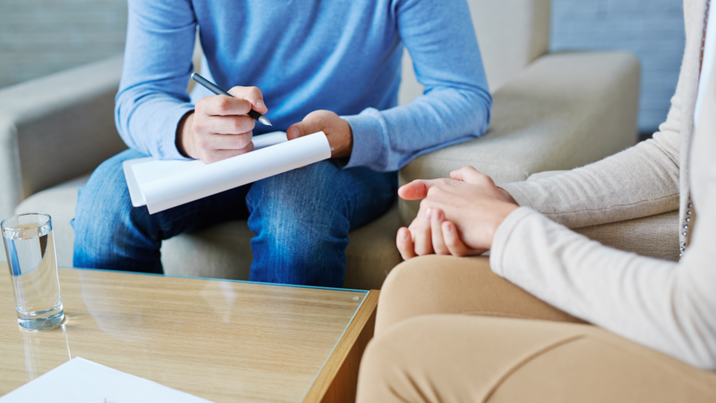 A therapist or counselor taking notes while talking to a patient in a therapy session, sitting on a sofa with a glass of water on the table.
