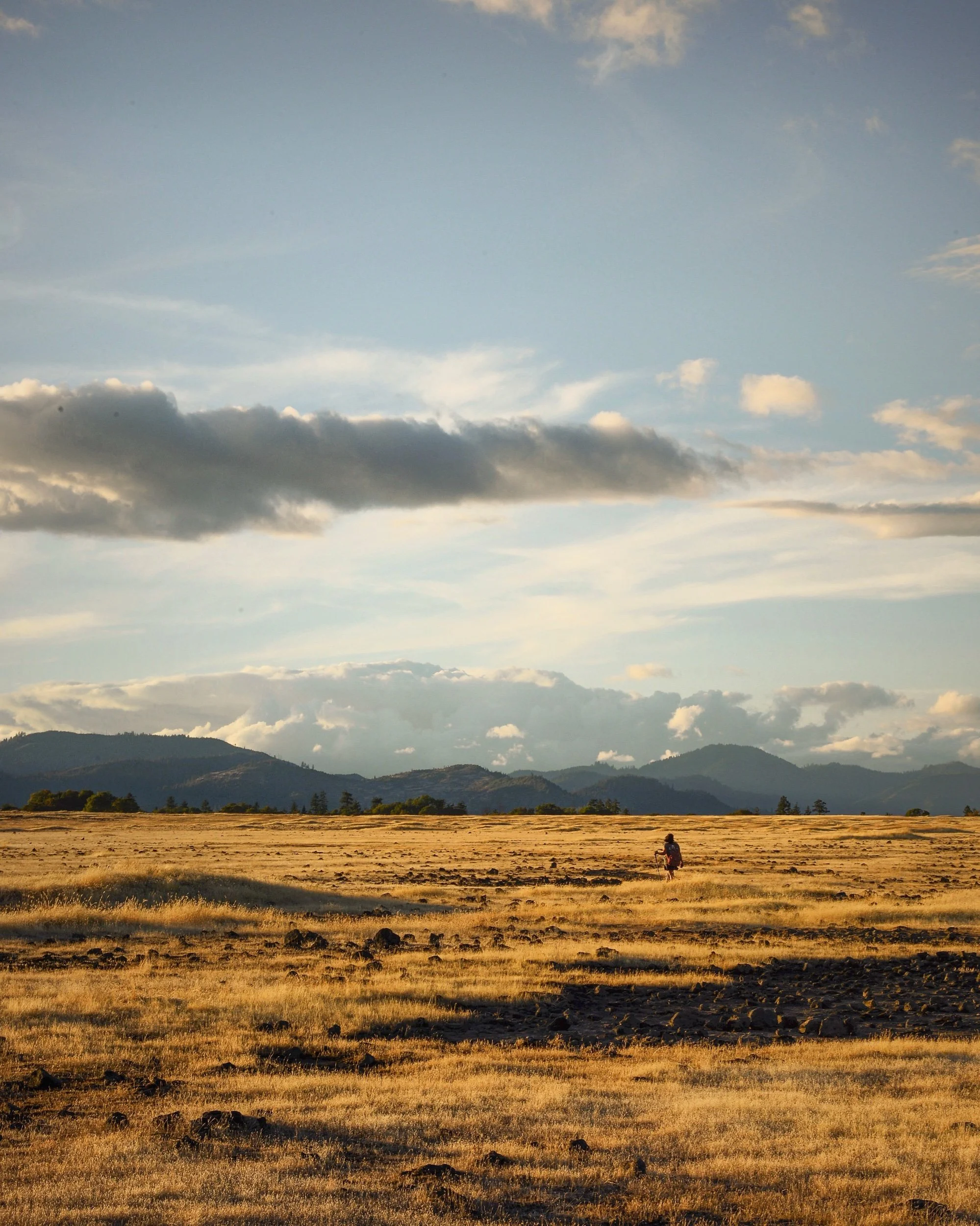 A person walking across a large, open field at sunset with mountains in the background and clouds in the sky.
