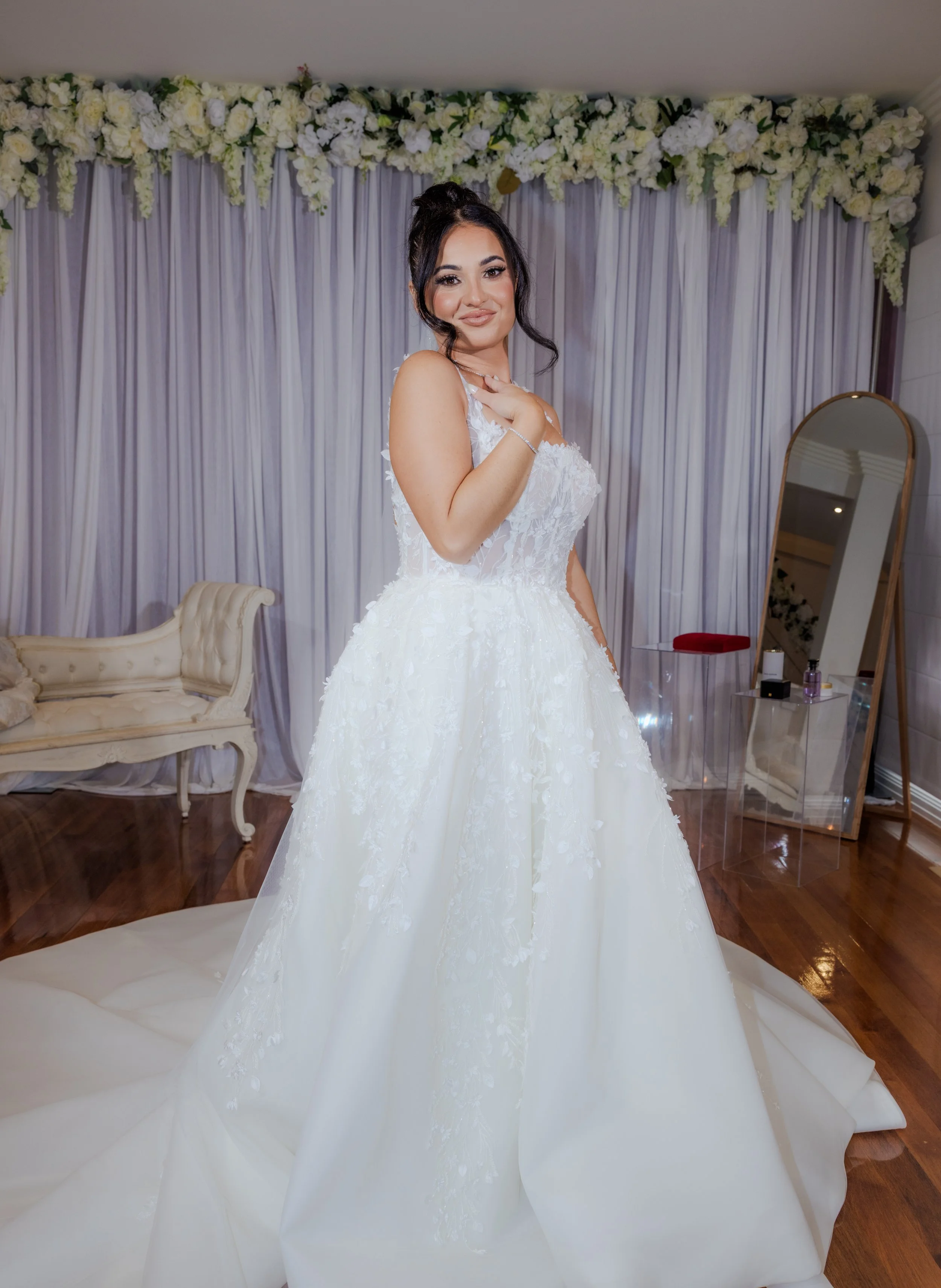 Brunette bride in a white wedding gown standing in a decorated room with floral arrangements and a mirror.