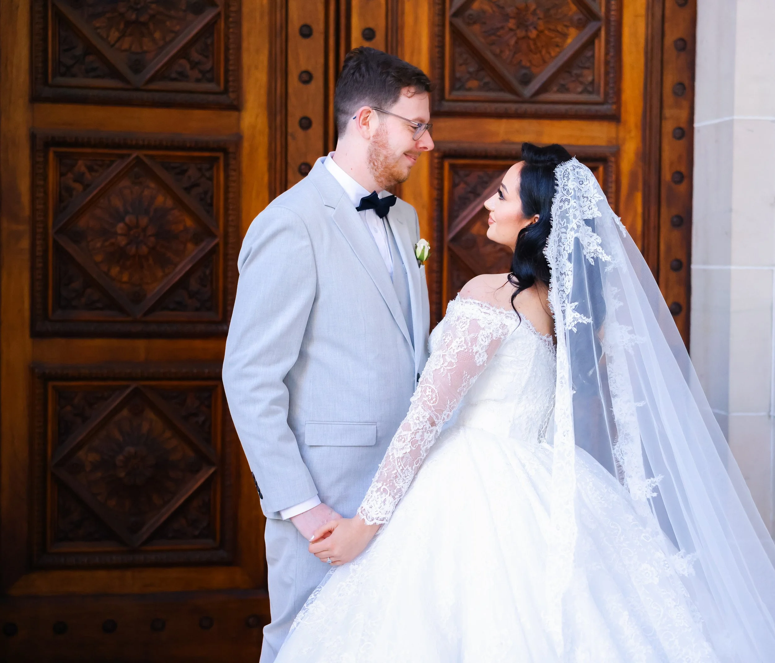 A newlywed couple holding hands in front of a wooden door, dressed in wedding attire, gazing at each other.