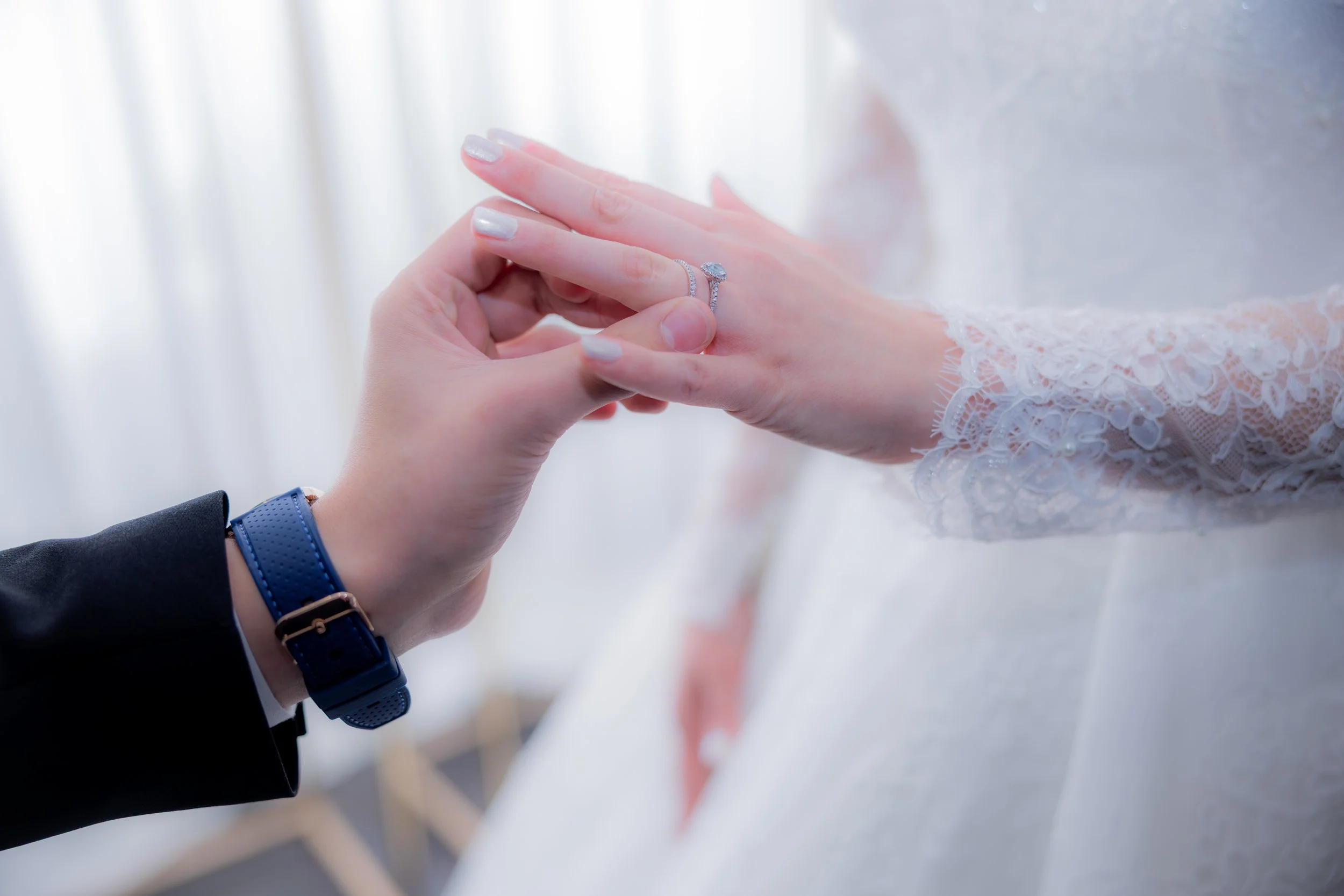 A close-up of a bride and groom during a wedding ceremony, showcasing the groom placing a ring on the bride's finger, with lace sleeves visible on her dress.