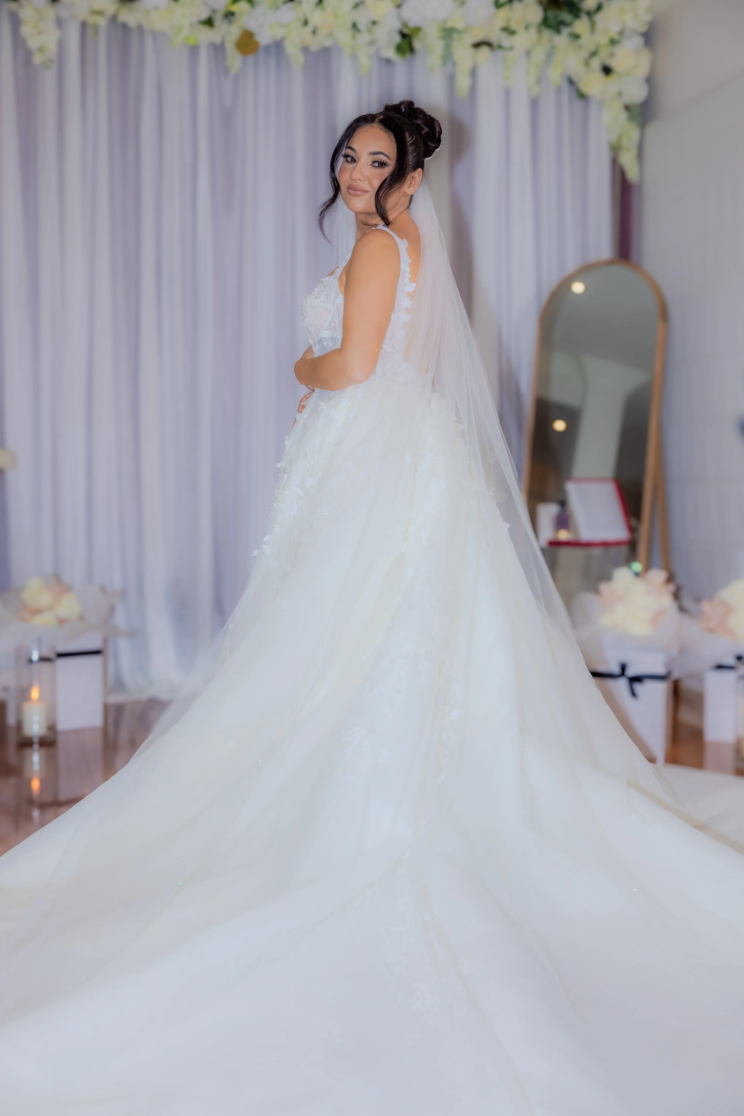 Bride in a white wedding gown and veil standing in a decorated room, smiling at the camera.