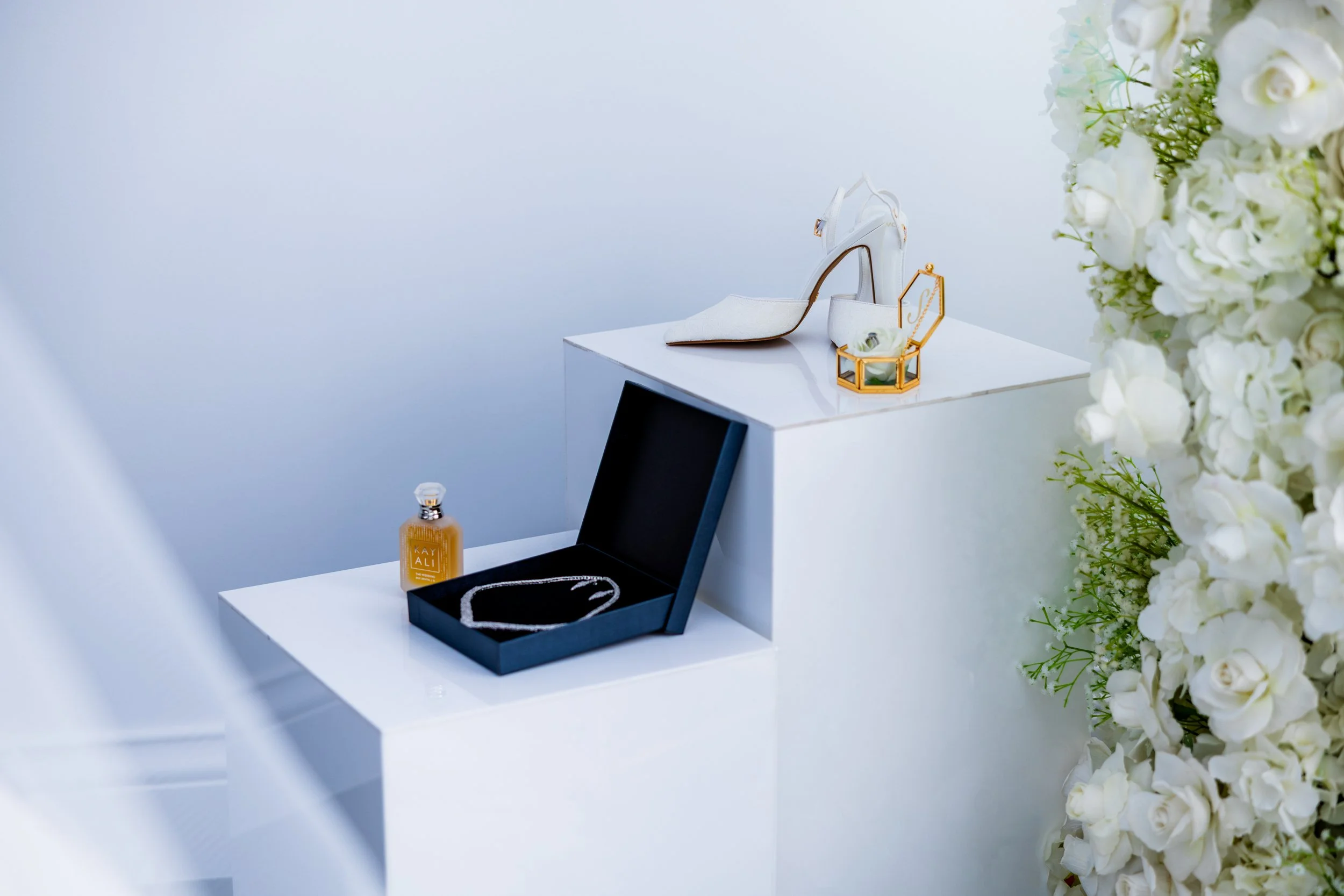 Display of jewelry and accessories on white pedestals, including a ring in a gold ring box, a necklace in an open black box, and a high-heeled white shoe with a gold heel, with white flowers on the right side.