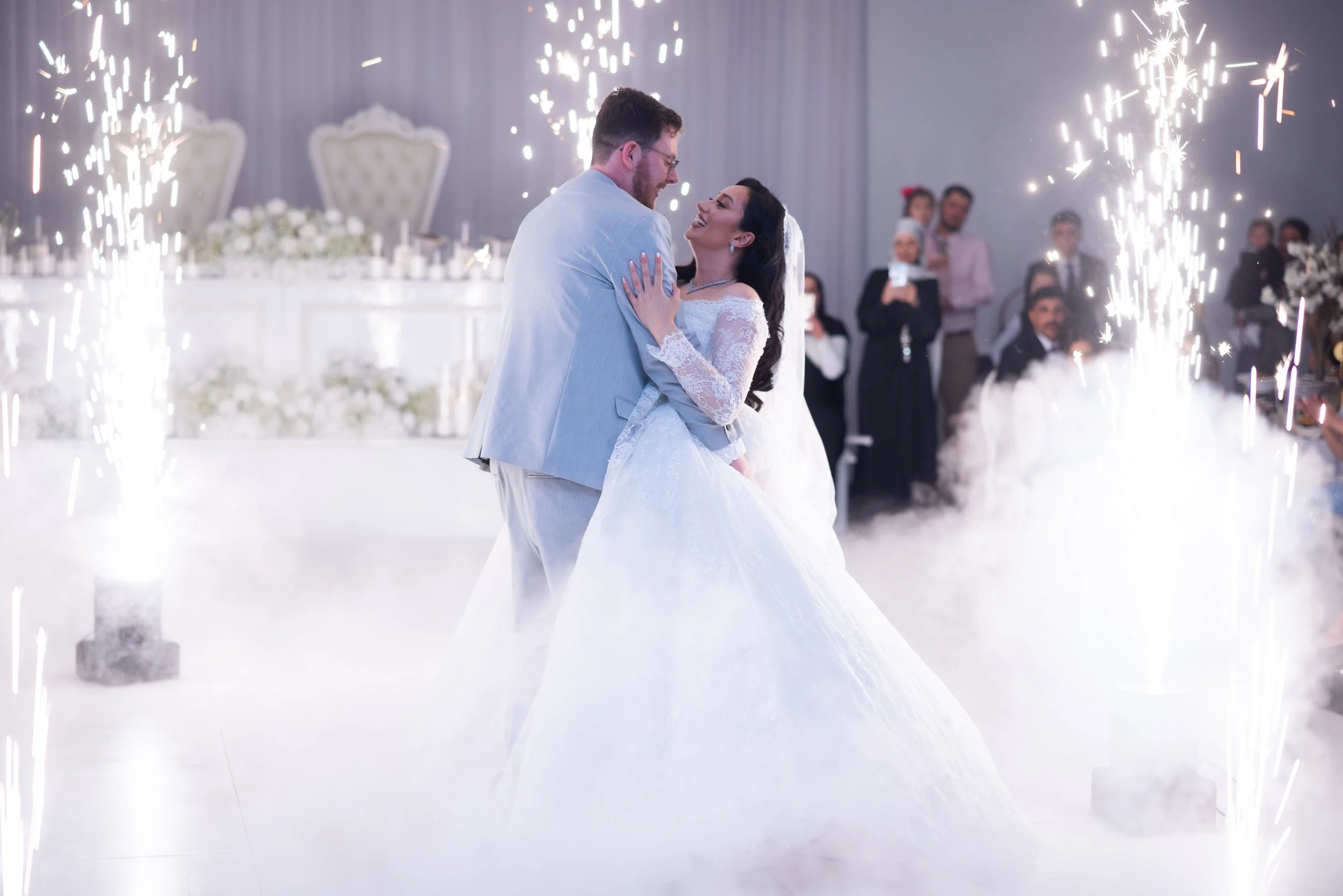 A newlywed couple dancing at their wedding reception surrounded by smoke and fireworks, with guests watching in the background.