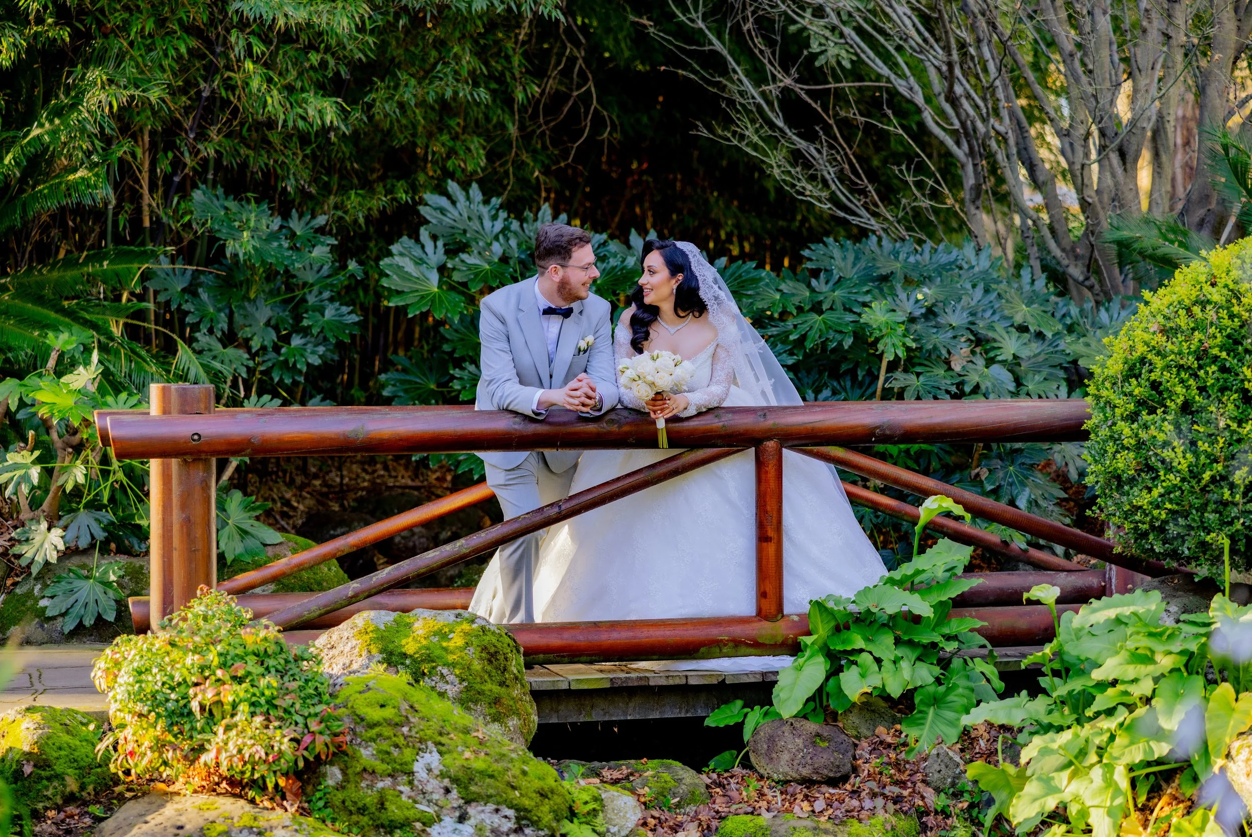 A bride and groom standing on a wooden bridge surrounded by lush green plants, smiling and holding hands, with the bride holding a bouquet of white flowers.