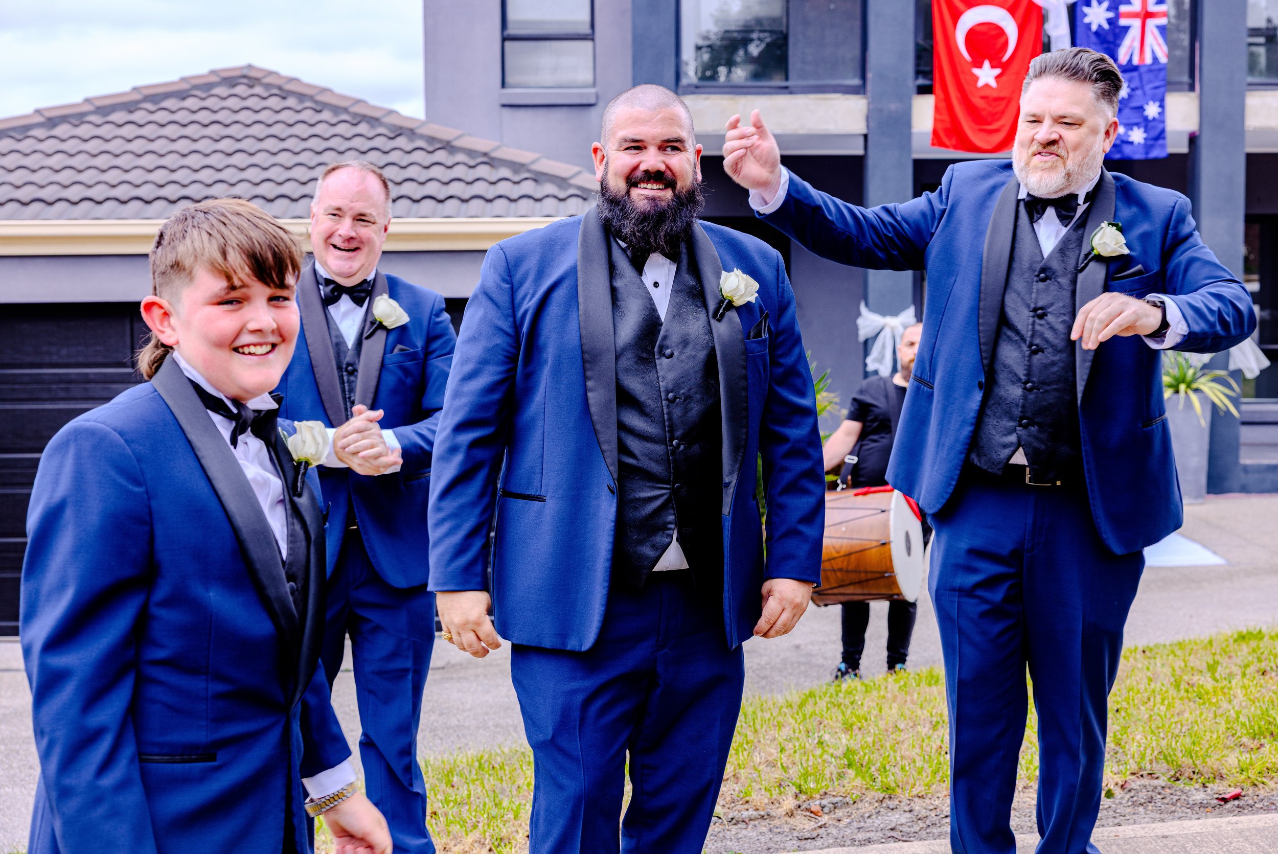 Group of men and a boy dressed in blue suits at a wedding, smiling and celebrating outdoors with flags in the background.