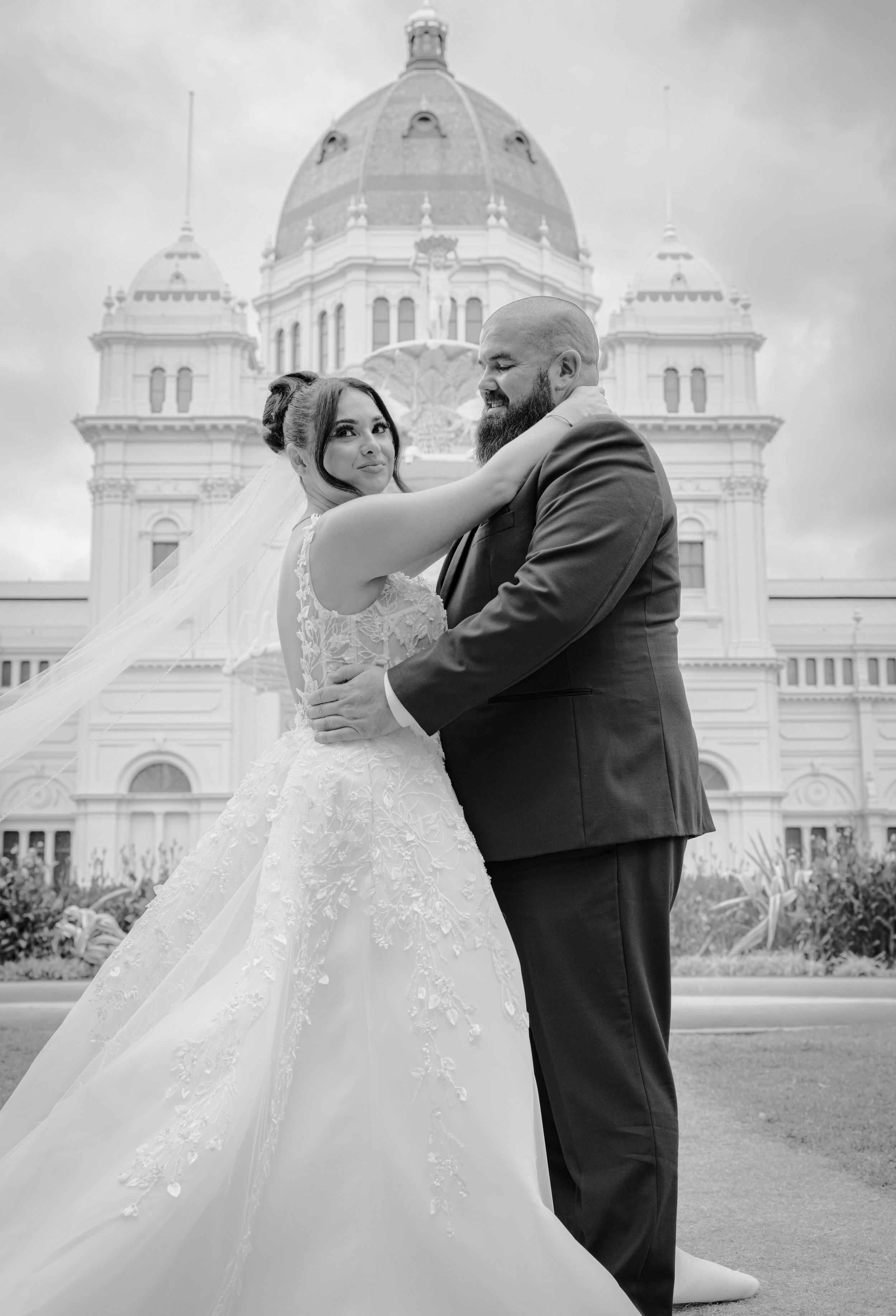 Black and white photo of a bride and groom hugging outside a large historic building with a dome and towers.