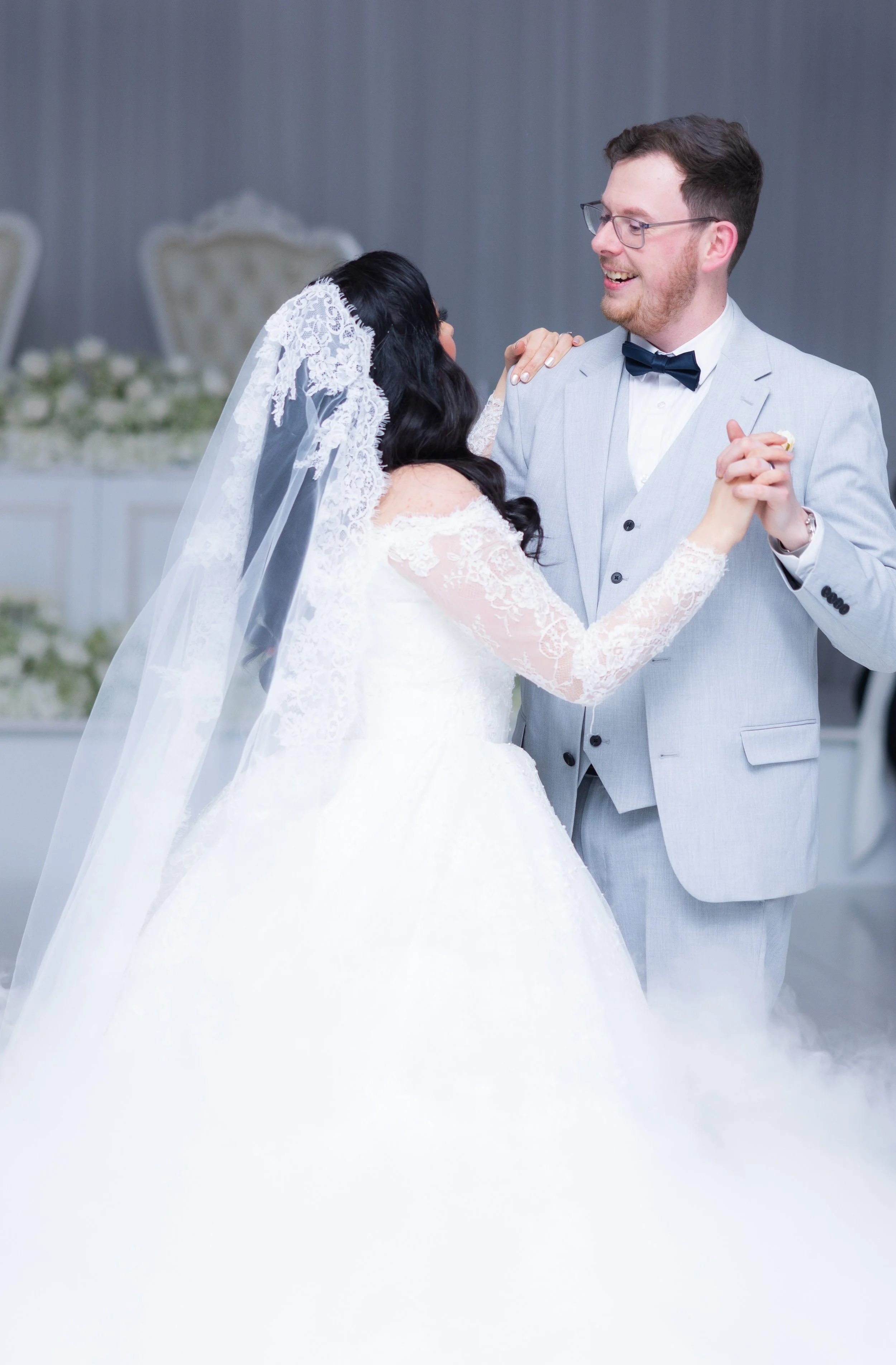 A bride and groom share a dance at their wedding reception, with the bride in a white lace wedding gown and veil, and the groom in a light gray suit with a black bow tie, in a decorated indoor venue.