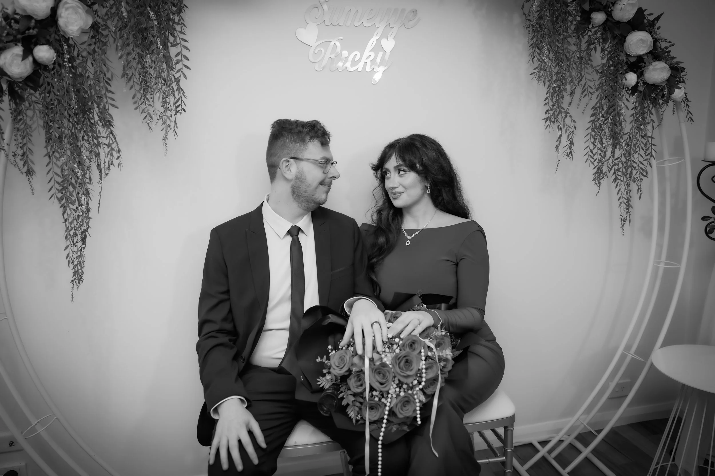 A couple sitting on a bench, the man in a suit and the woman in a dress, holding a bouquet of flowers, looking at each other warmly, with floral decorations and a sign that says 'Sumeyye & Rıfkı' above them.