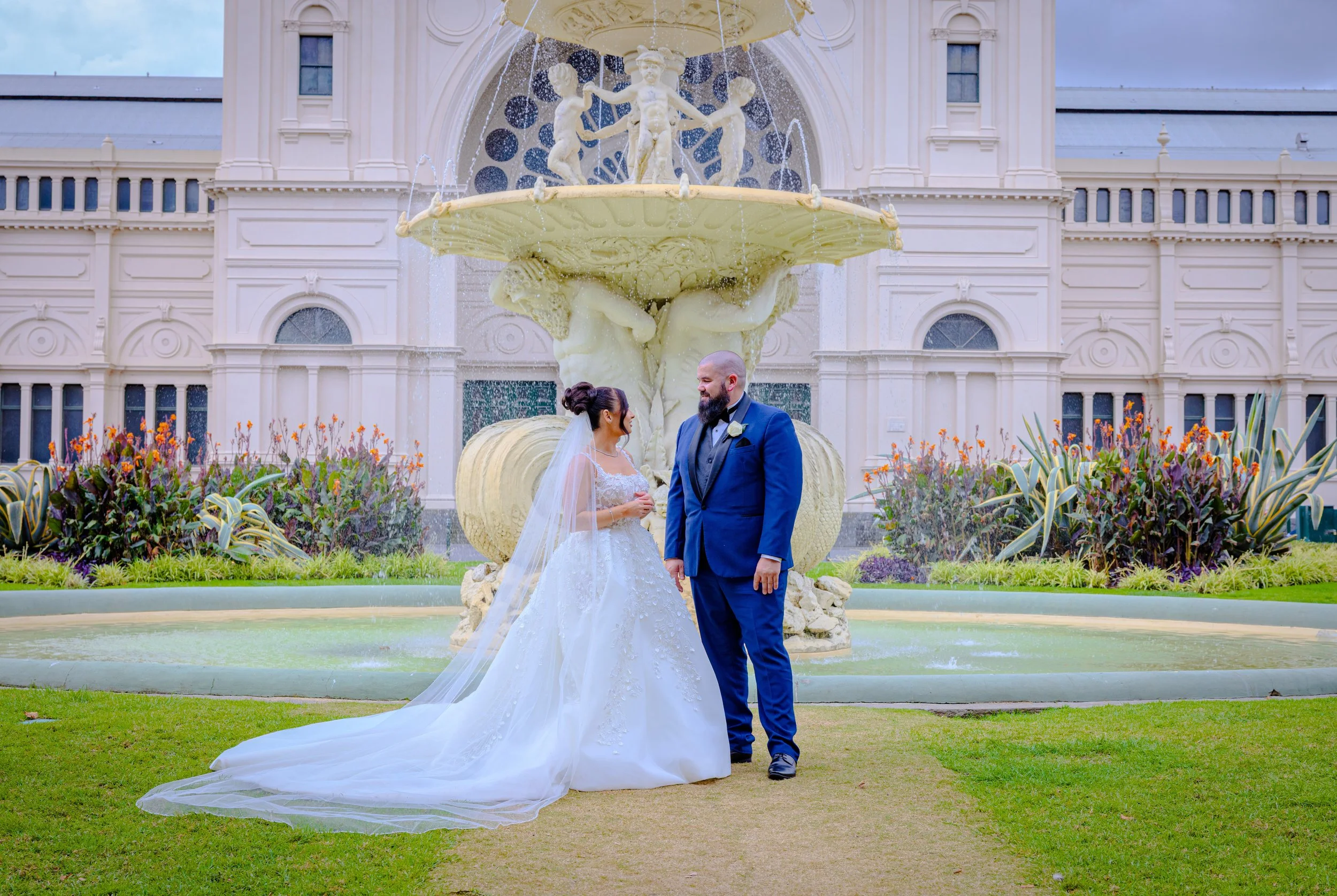 A bride and groom standing in front of a large decorative fountain with statues, surrounded by flowers and greenery, during their wedding ceremony.