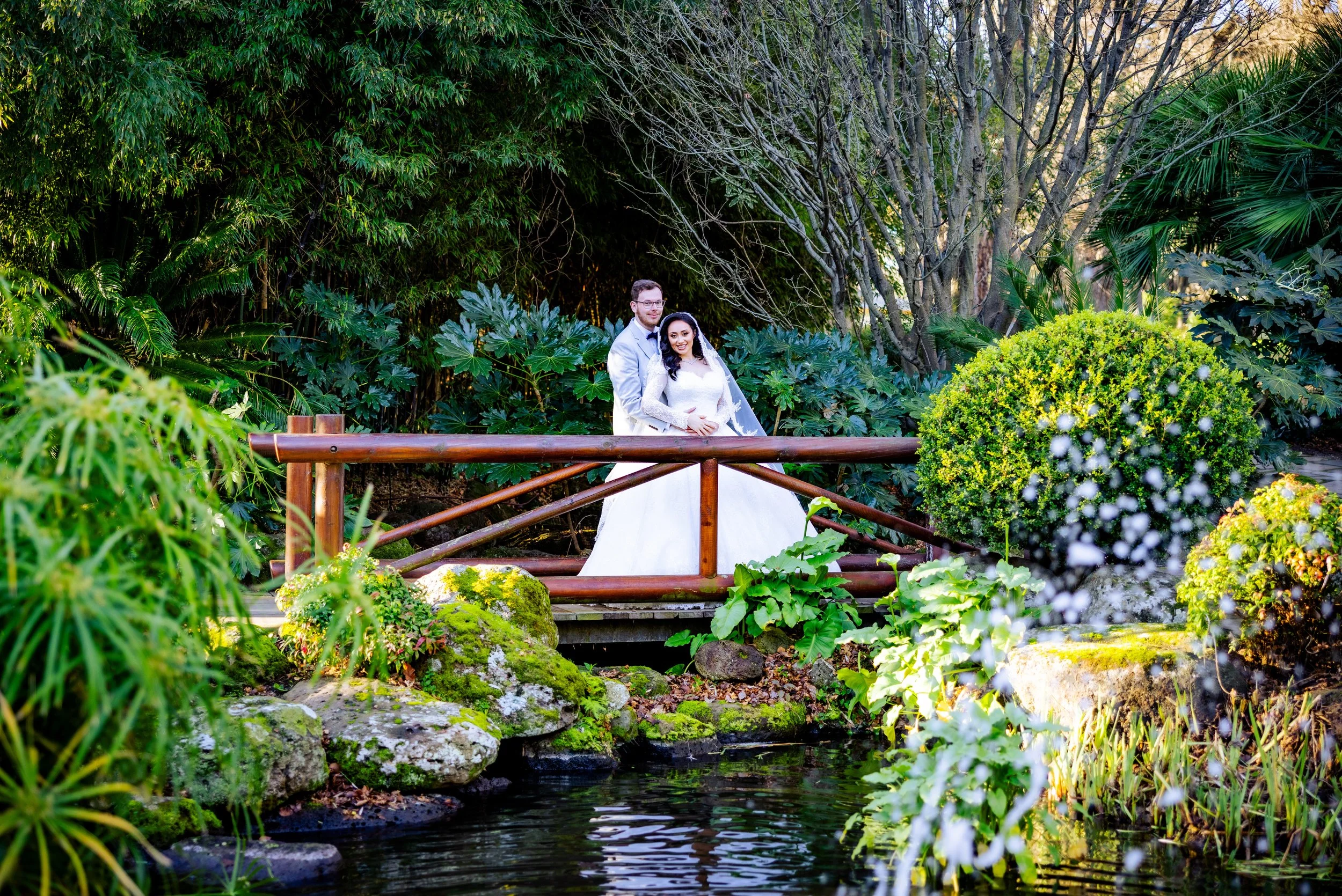 A newlywed couple standing on a small wooden bridge over a pond in a lush garden with green foliage and rocky landscaping.