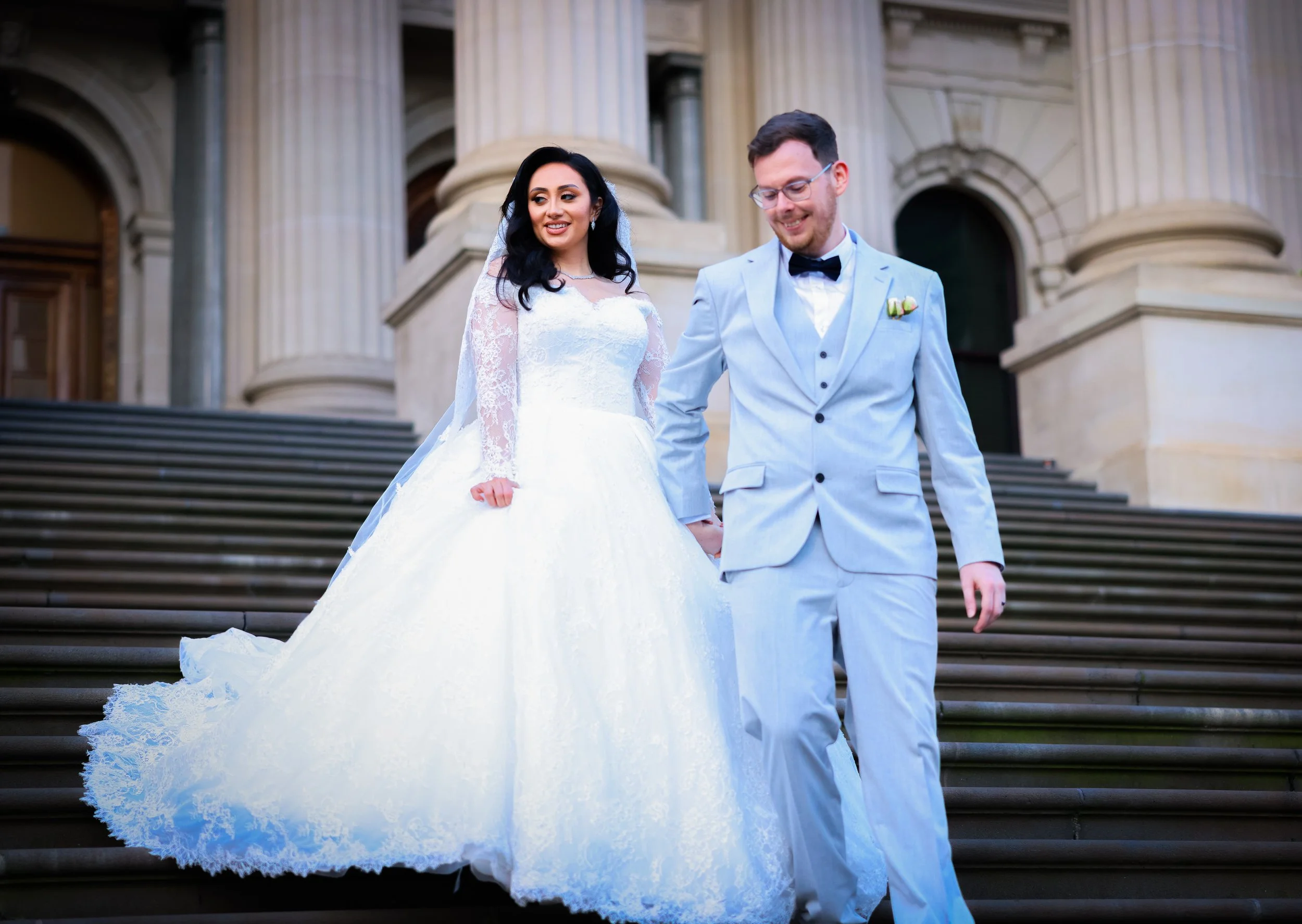 A bride and groom holding hands and walking down the steps of a historic building with large columns. The bride is in a white wedding dress with lace details, and the groom is in a light blue suit with a black bow tie.
