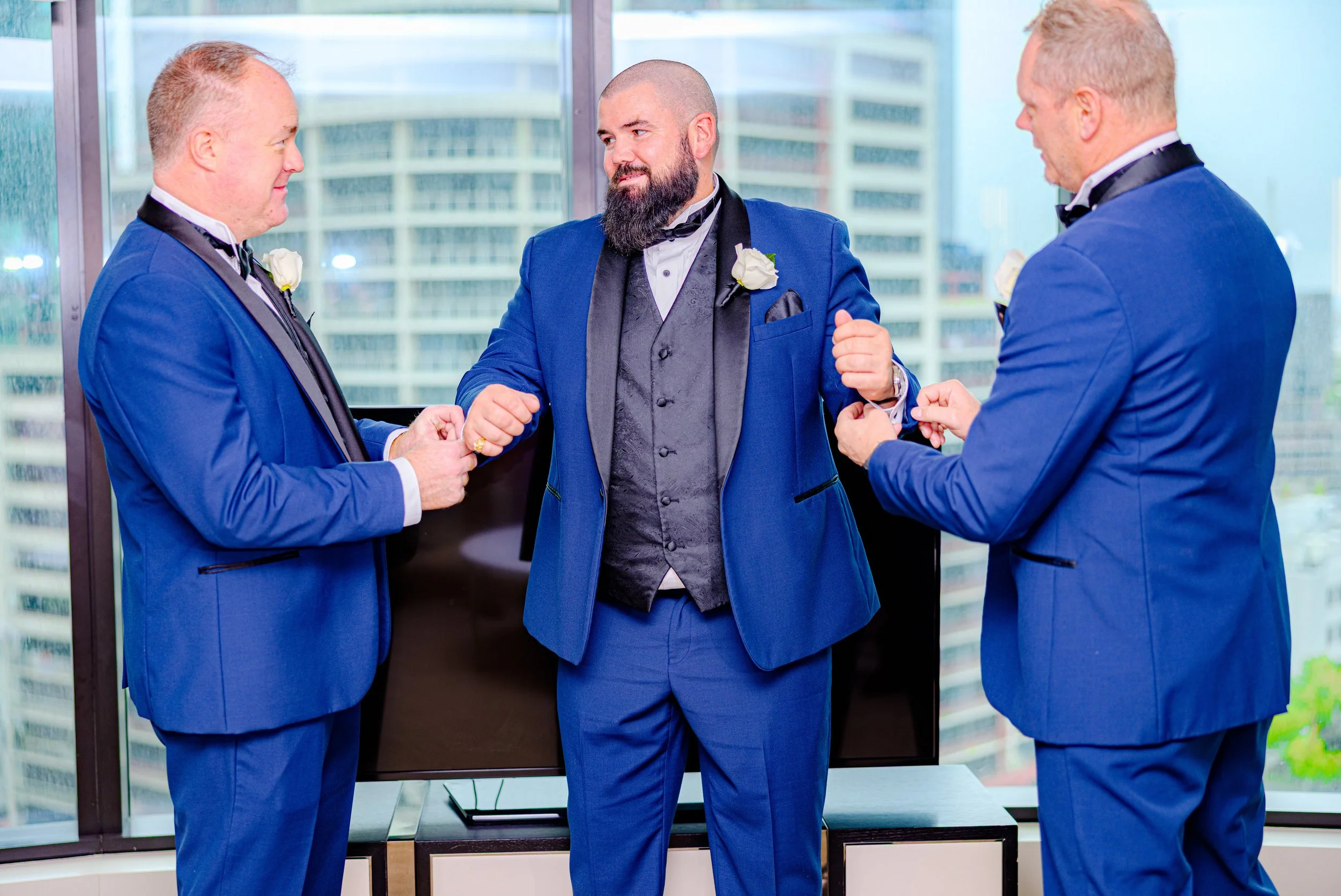 Three men in blue tuxedos, one with a beard, standing indoors in front of large windows with city buildings in the background, engaging in conversation and adjusting their cufflinks.