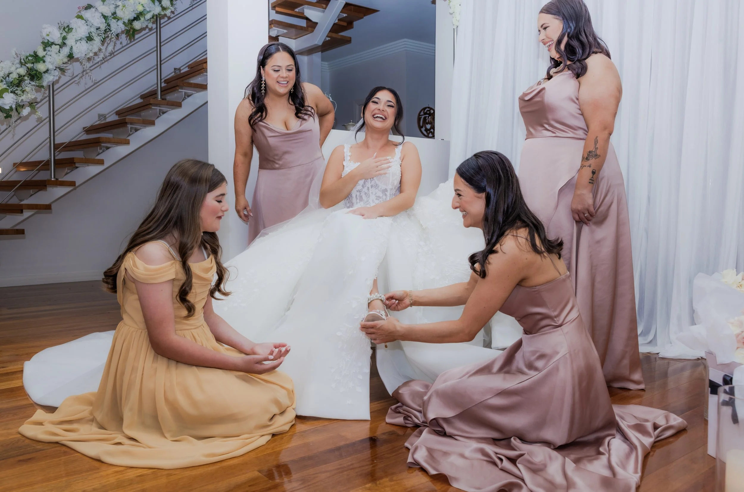 A bride sitting on a white sheet with five women around her, one helping her put on a shoe, smiling and laughing while getting ready for her wedding in a well-lit room decorated with white drapes and flowers.