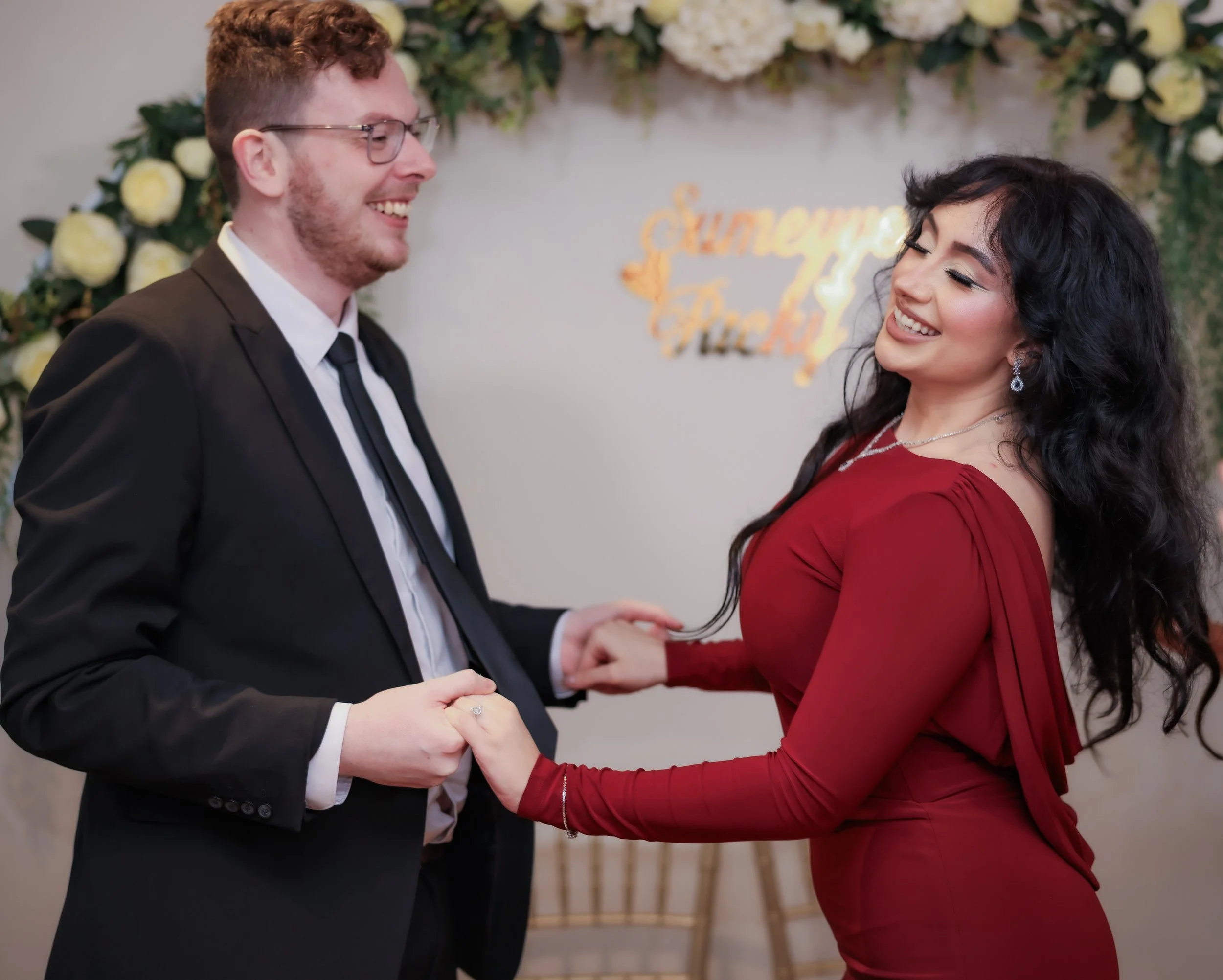 A couple dancing and smiling at their wedding reception, with a floral backdrop and a sign that says 'Happily Ever After'.