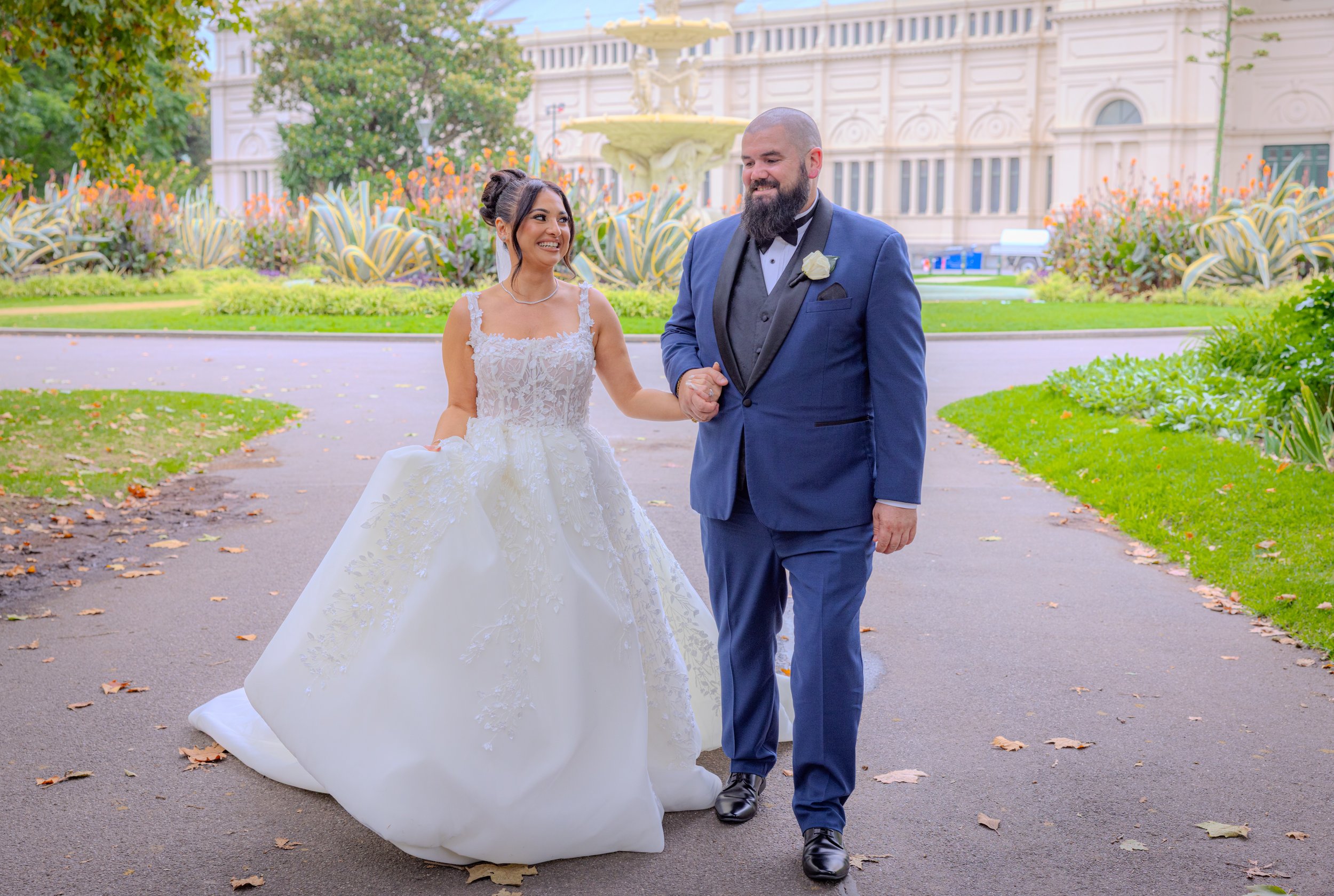 A newlywed couple walking together outdoors in a park, with the bride in a white wedding gown and the groom in a blue tuxedo, holding hands and smiling.