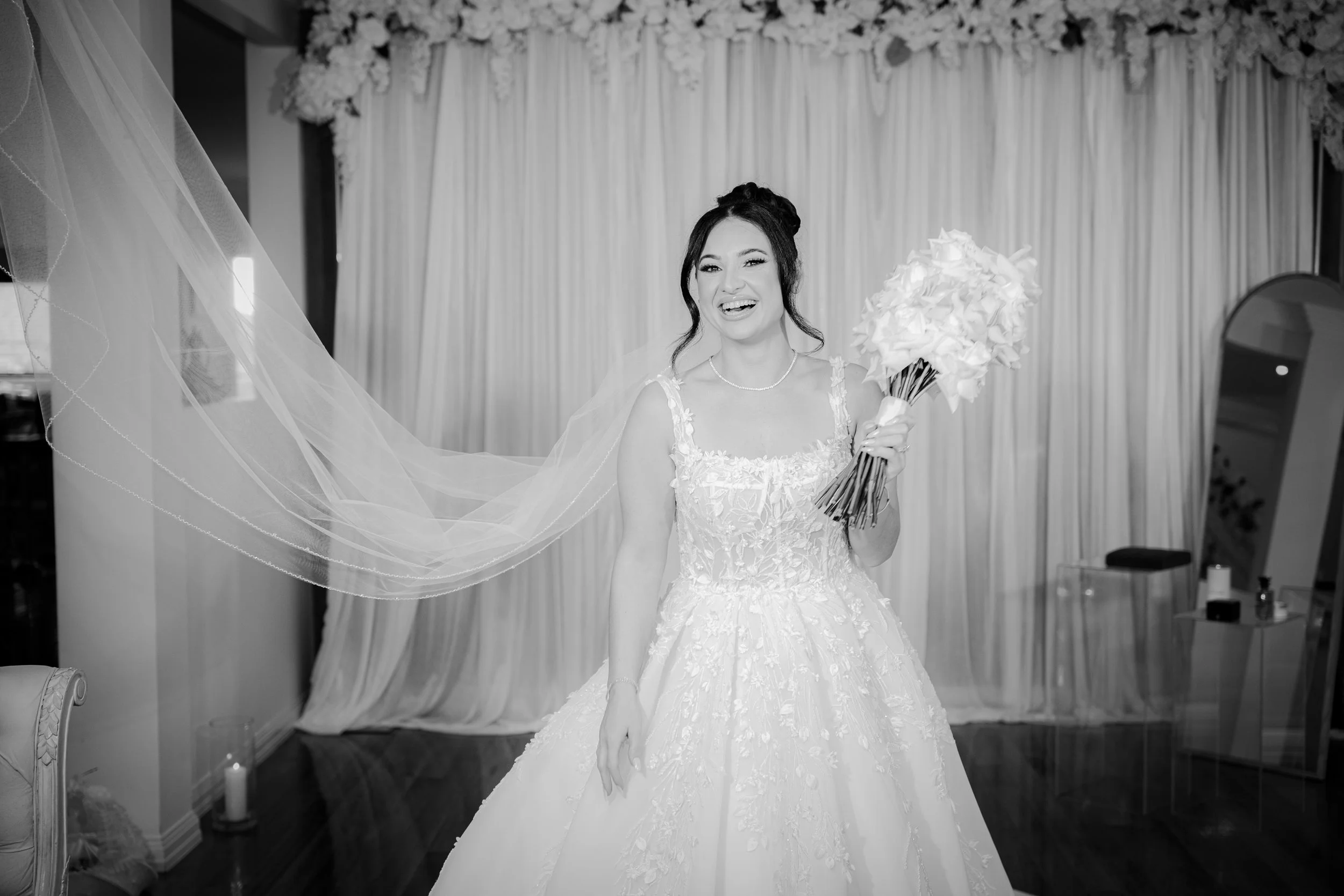Black and white photo of a bride smiling, holding a bouquet, in a room with curtain backdrop and floral decorations.