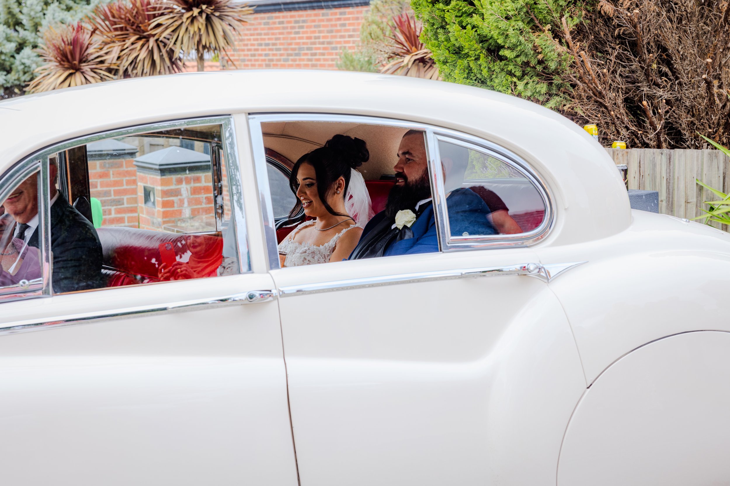 Bride and groom sitting in a vintage white car, smiling and looking at each other, during a wedding celebration.
