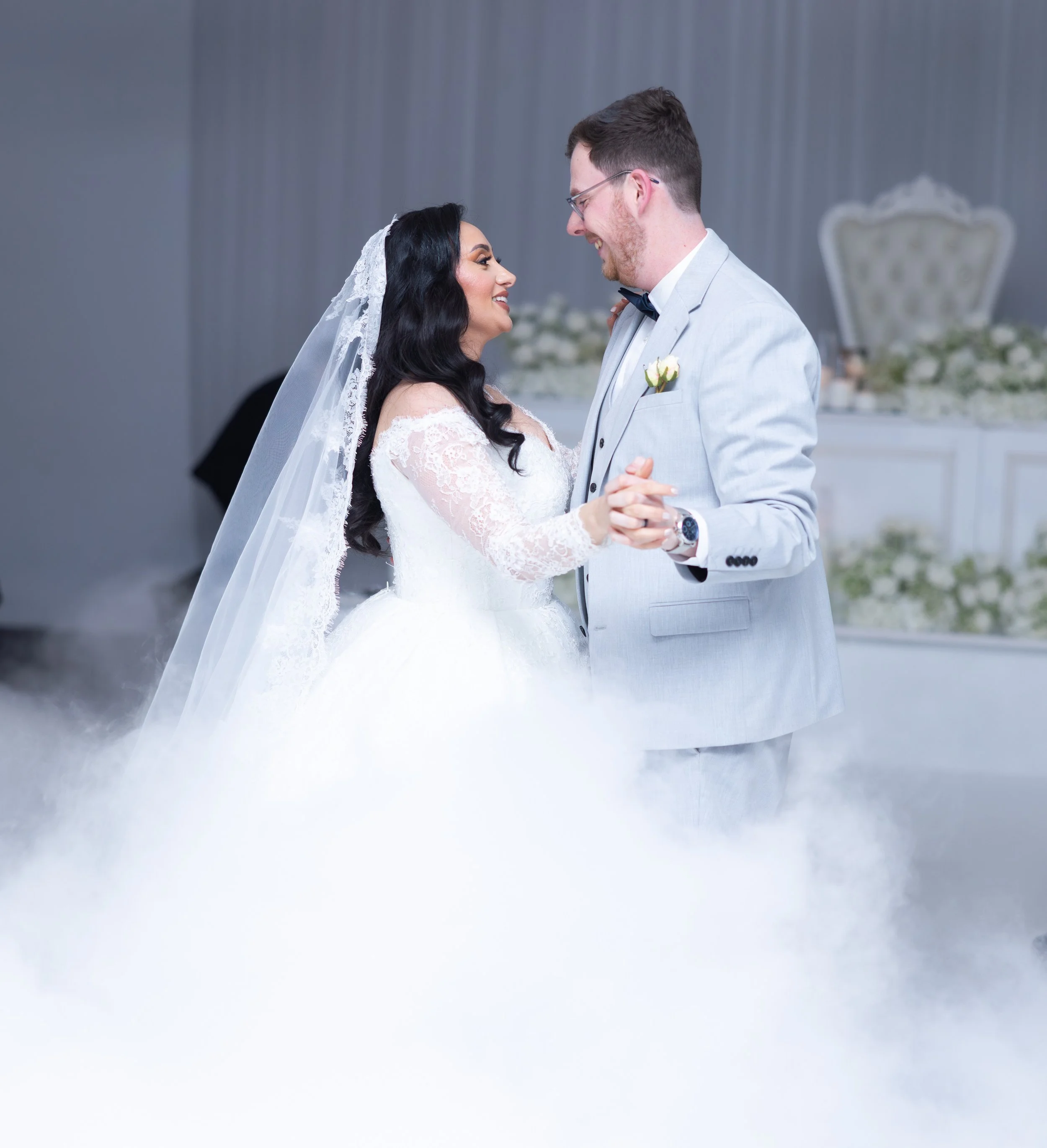 A bride and groom dance together at their wedding, smiling at each other, with a foggy dance floor and elegant background.