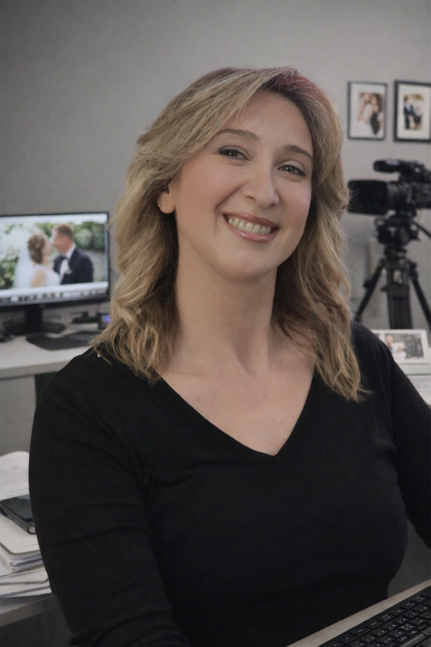 A woman with blonde hair smiling at the camera in an office setting, with a photo of a wedding on her computer screen and a professional video camera in the background.