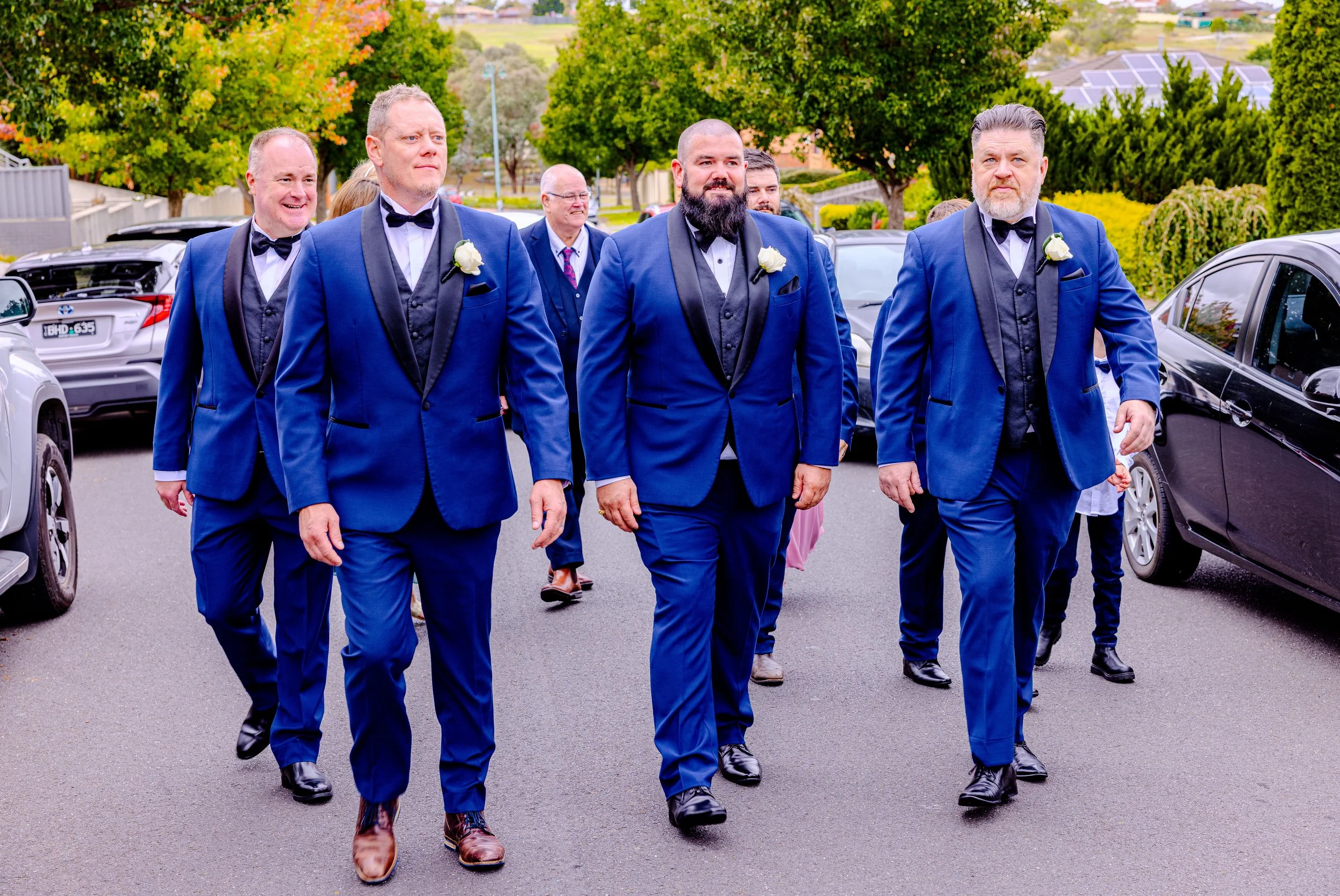 A group of men in blue tuxedos with black lapels and white boutonnieres walking together outdoors on a paved driveway.