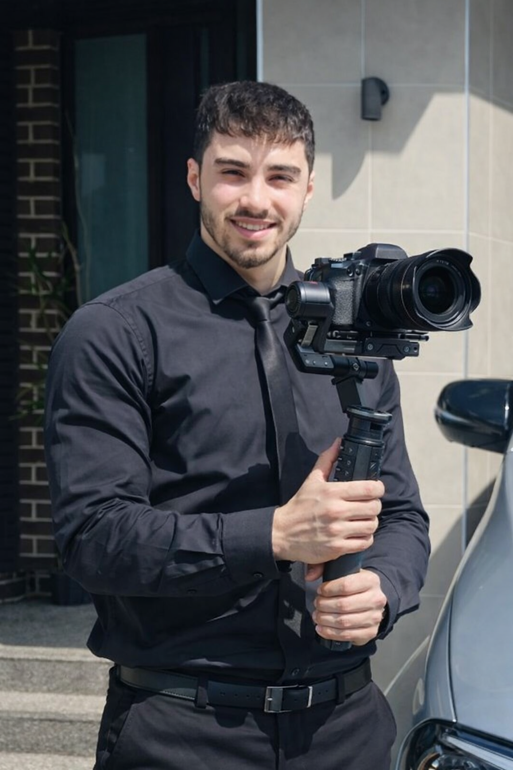 A man dressed in black holding a stabilizer with a professional camera attached, standing outside near a building.