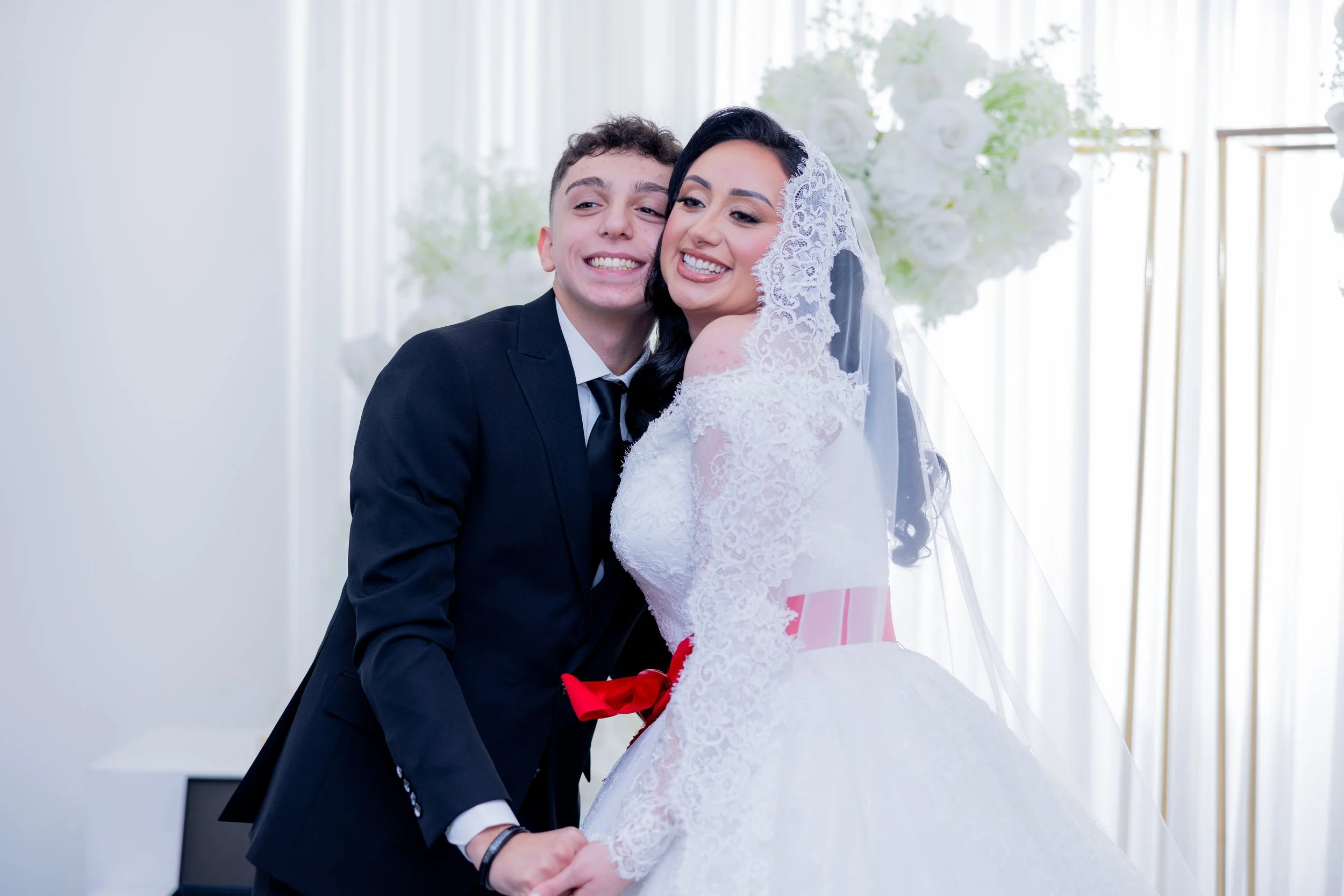 A newlywed couple, a man in a black suit and a woman in a white wedding gown with lace details and a veil, smiling and holding hands during their wedding celebration.