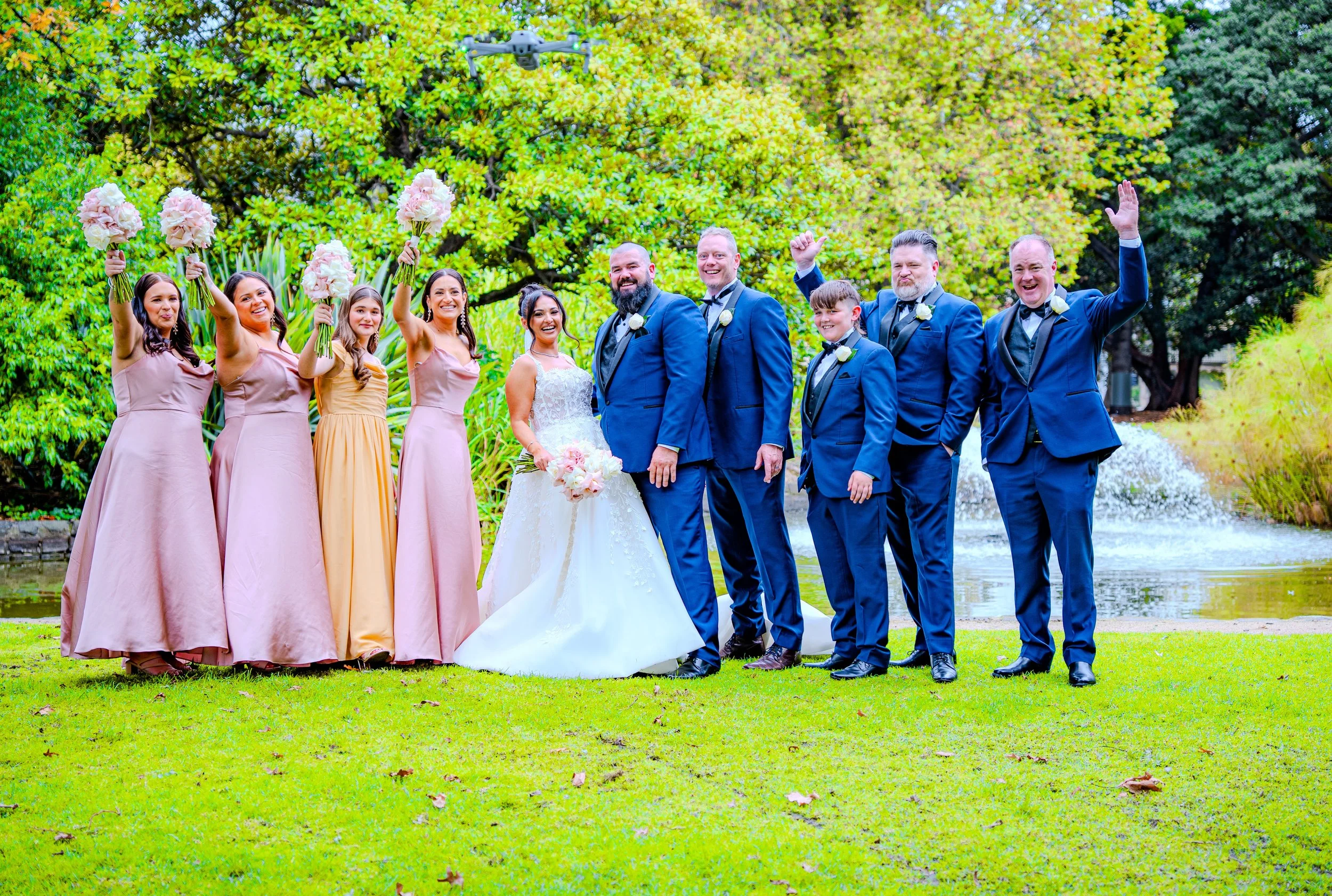 Wedding party standing on grass near a pond with fountain. The bride in a white gown holds a bouquet, surrounded by bridesmaids in pastel dresses, and groomsmen in blue suits. Some are smiling and celebrating.