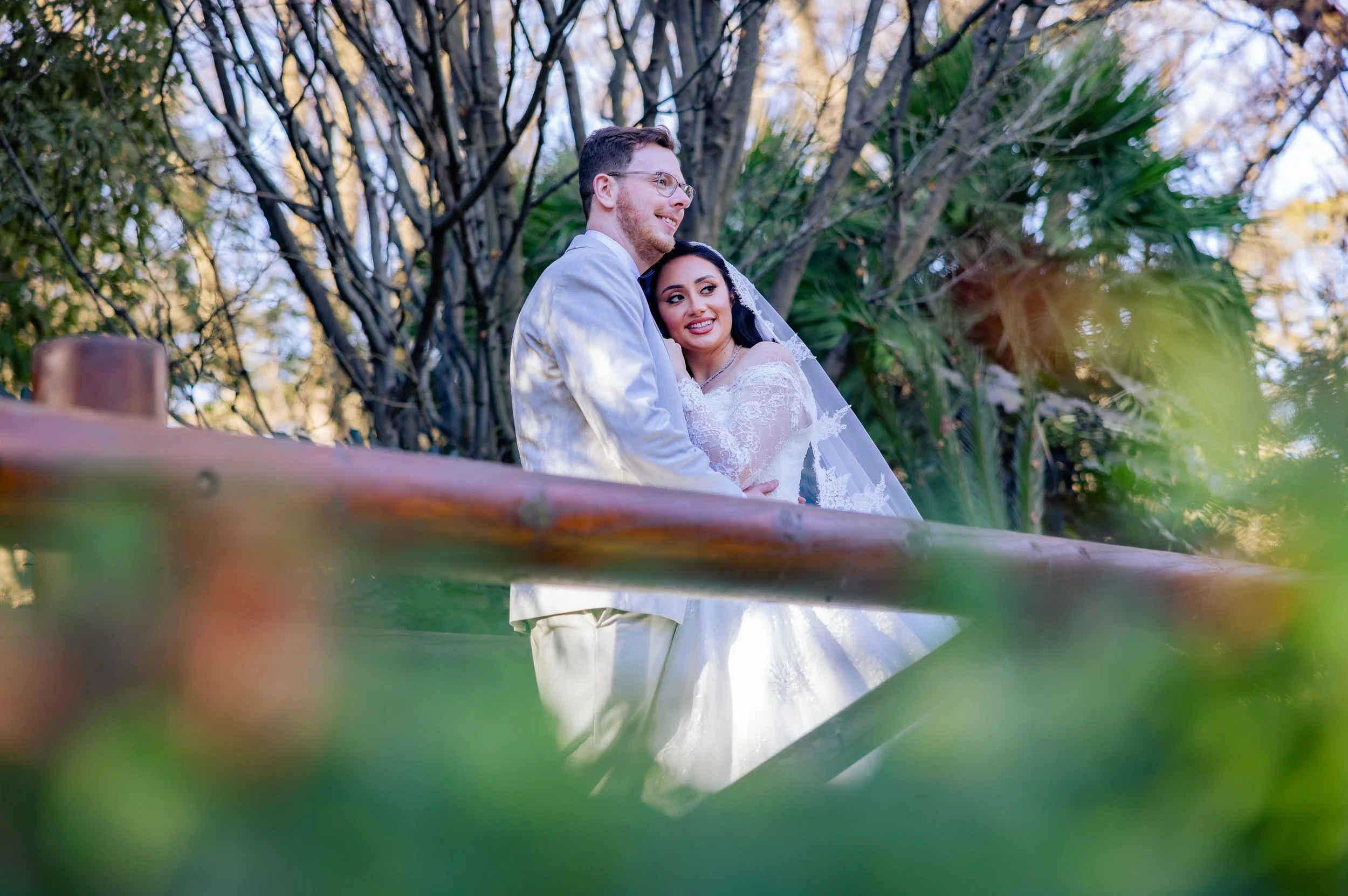 A newlywed couple dressed in wedding attire standing outdoors behind a wooden railing among trees and greenery, sharing a joyful moment.
