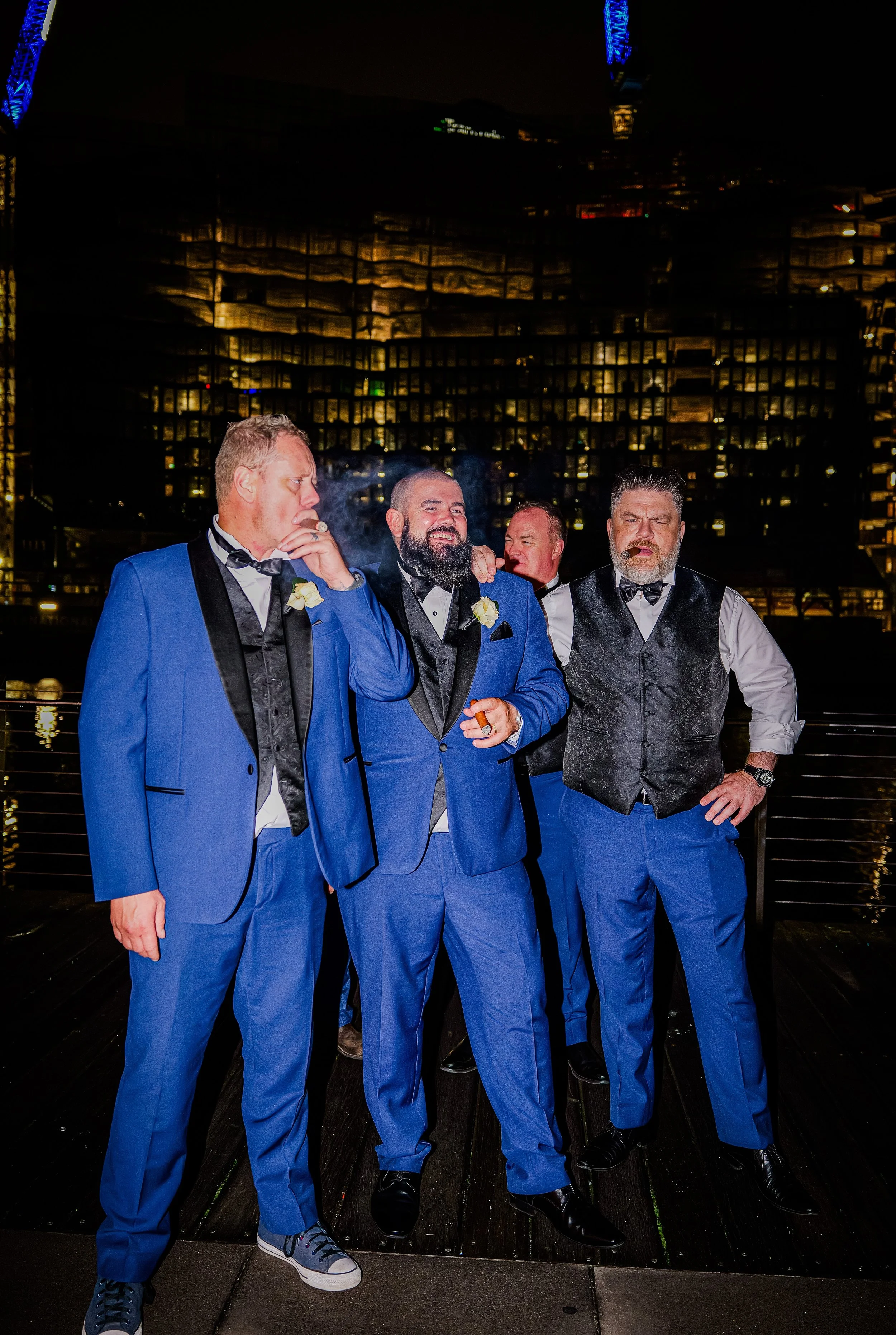 Four men dressed in tuxedos with blue pants and dark vests, standing at night on a rooftop with city lights in the background, smoking cigars, and enjoying themselves.