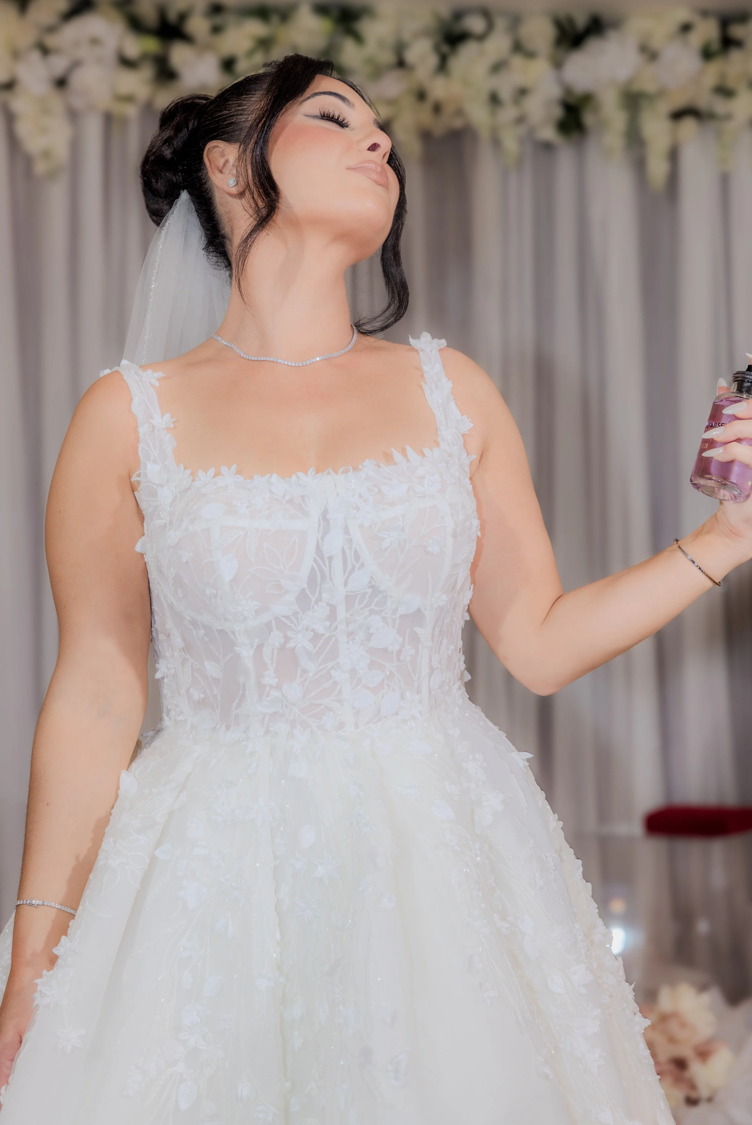 A bride in a white wedding dress with floral lace details, wearing a veil and jewelry, standing with her eyes closed and holding a perfume bottle.