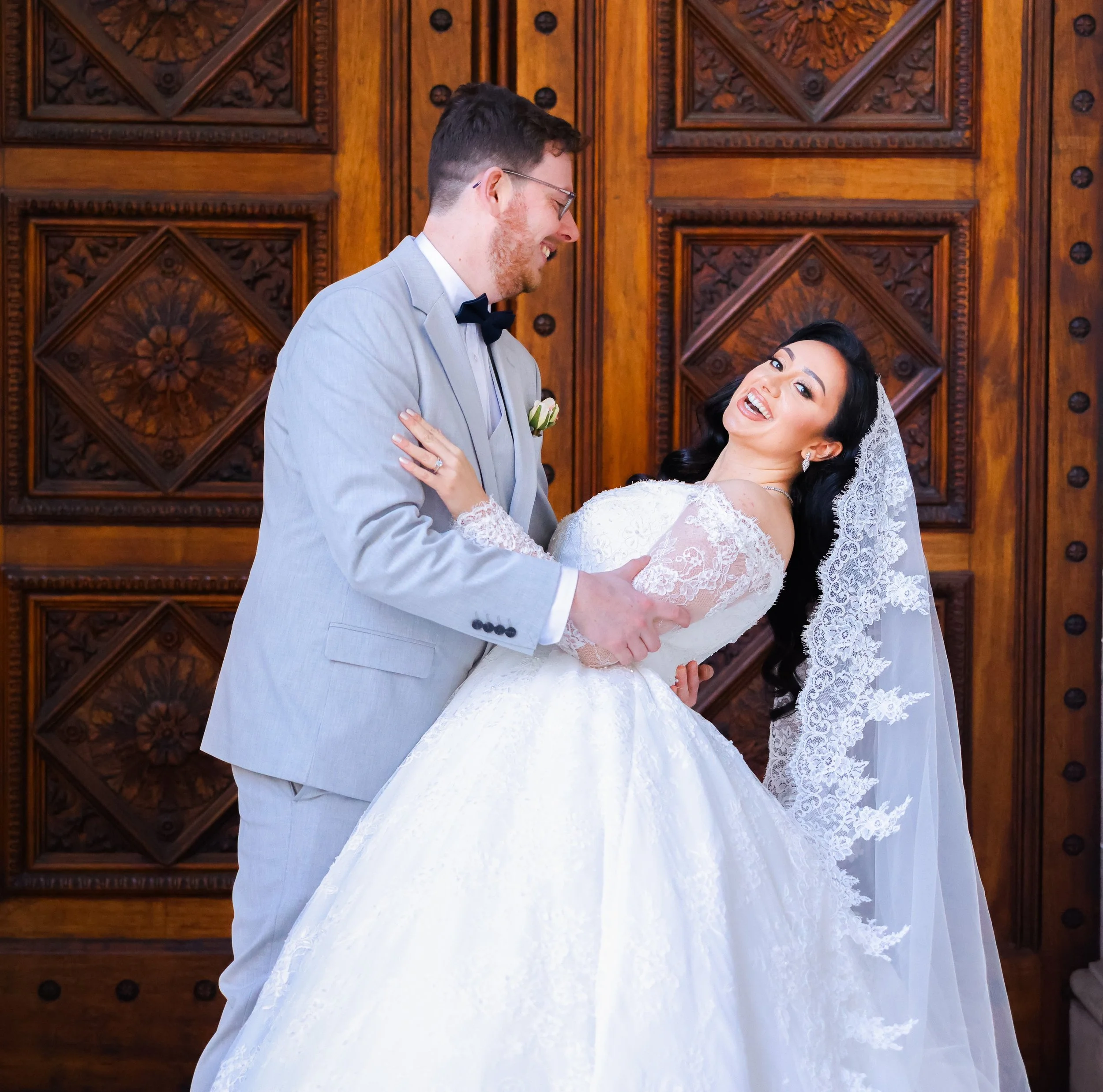 A newlywed couple shares a joyful moment, with the groom holding the bride in a dance pose against a richly carved wooden background.