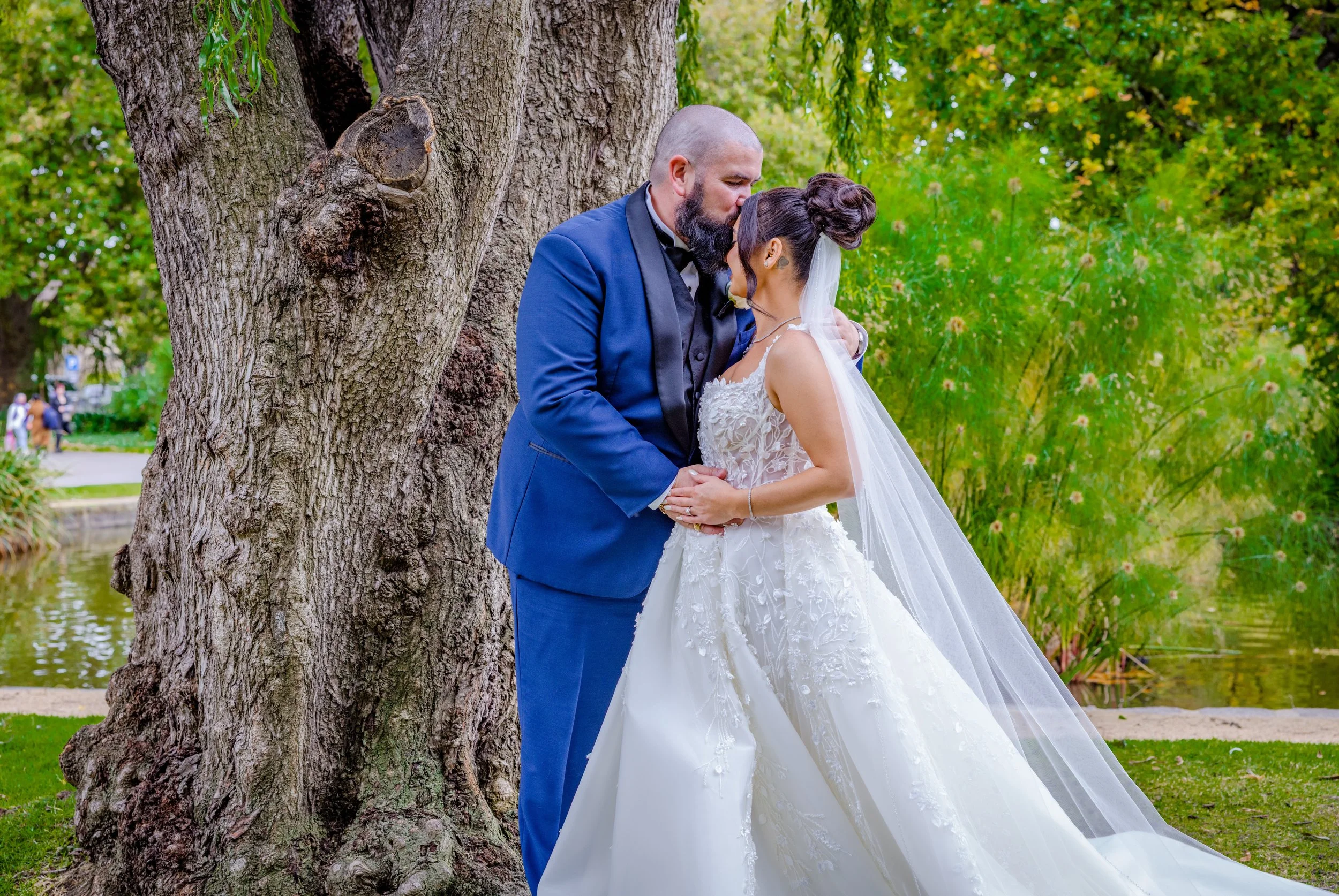 A bride and groom standing close together outdoors near a large tree, with the groom kissing the bride on her forehead. The bride is wearing a white wedding gown with lace details and a long veil, and the groom is dressed in a blue suit with a black 