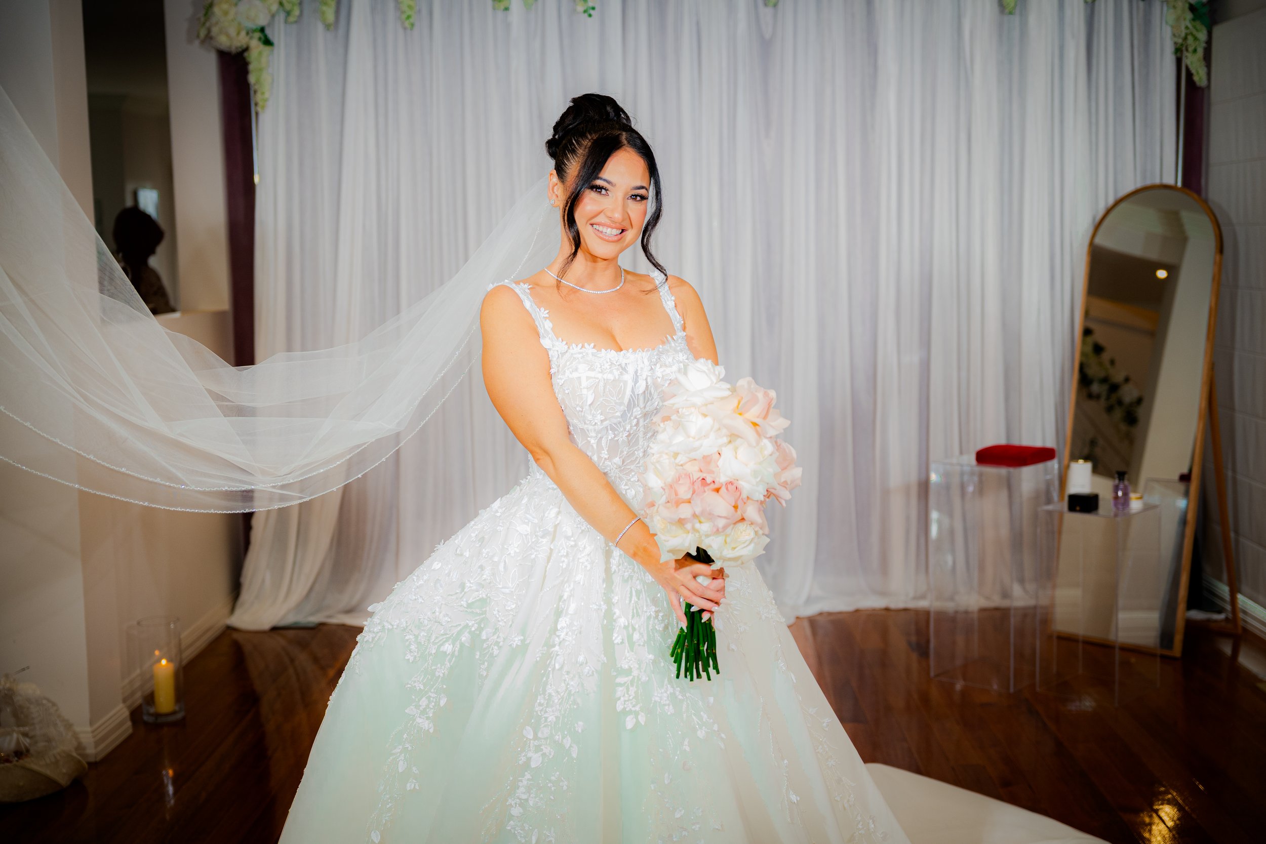 A bride in a white wedding gown holding a bouquet of white and blush pink flowers, standing in a decorated room with a white curtain backdrop, a mirror, and candles.