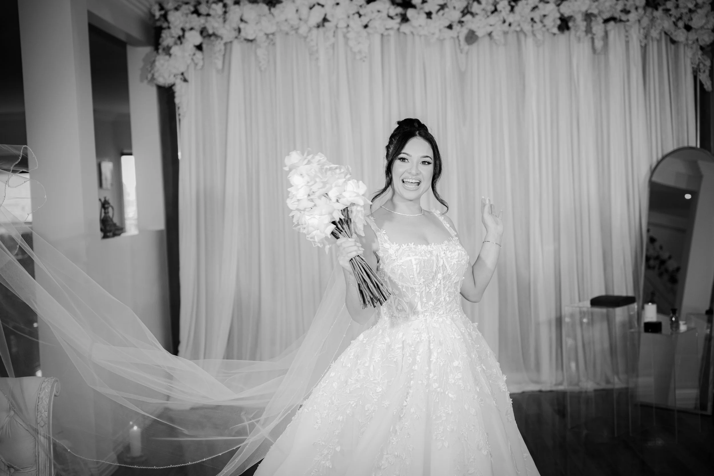 A woman in a wedding dress holding a bouquet of flowers, smiling and waving in front of a decorated backdrop.