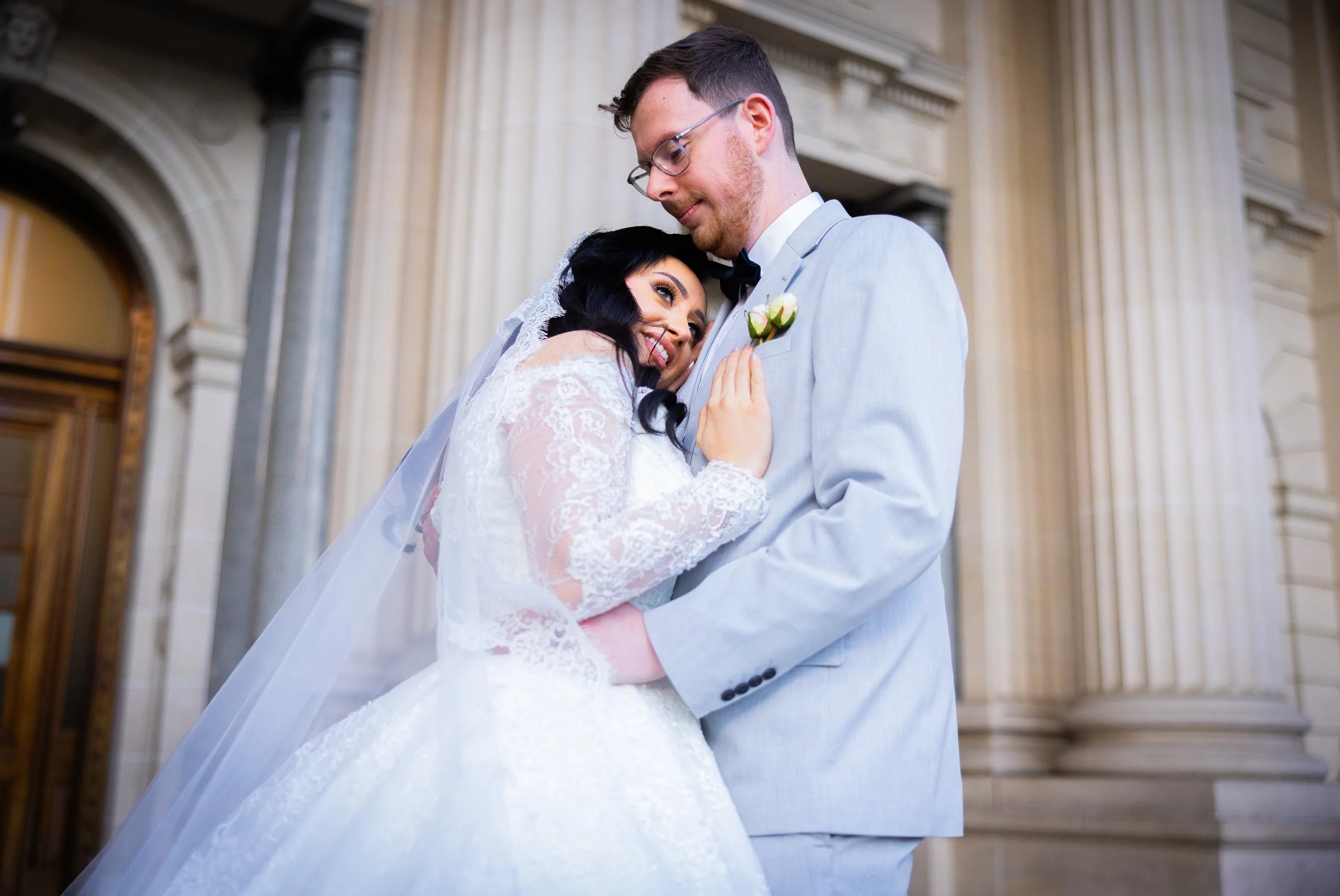 A bride and groom share an intimate moment on their wedding day inside a grand building with tall columns. The bride wears a white lace wedding dress and veil, while the groom wears a light gray suit with a boutonniere.