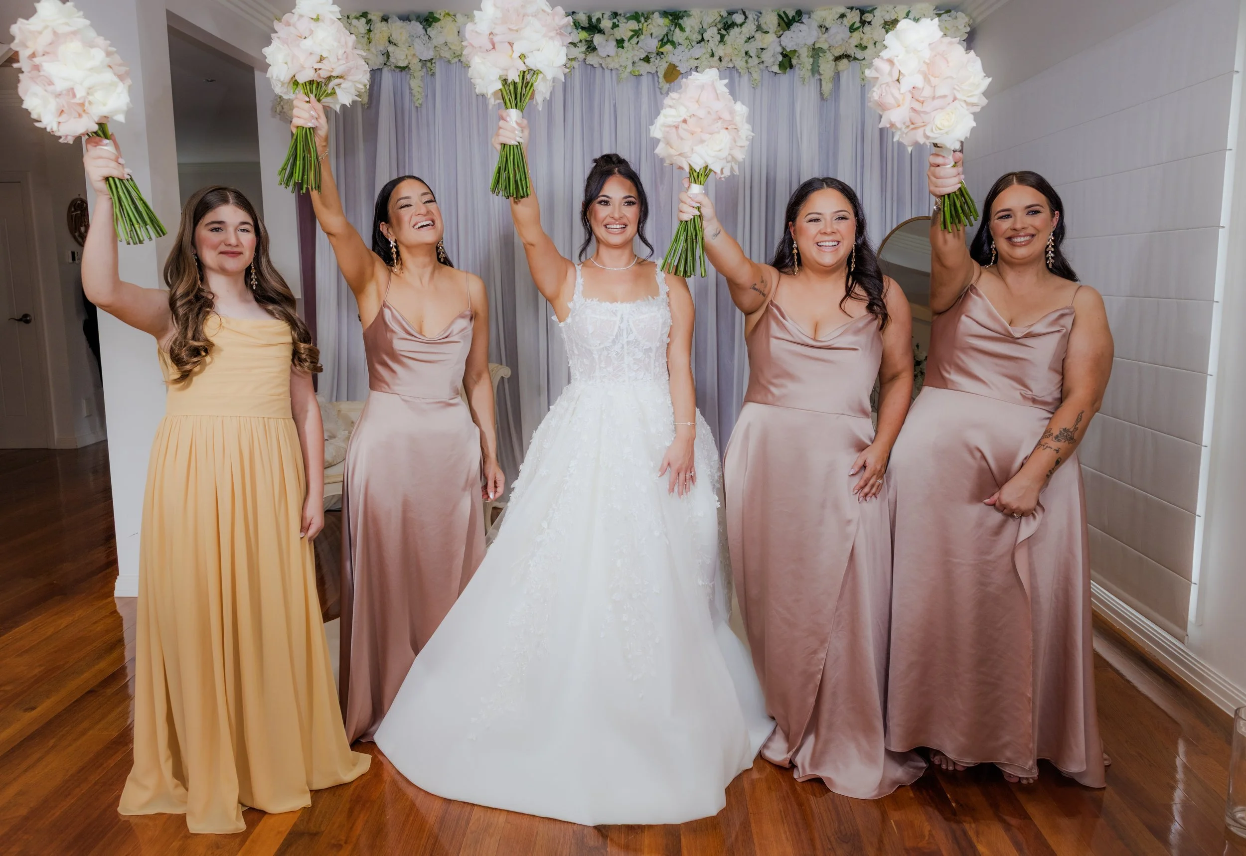 Bride in a white wedding dress holding a bouquet, surrounded by four bridesmaids in matching satin dresses, all smiling and raising their bouquets in celebration during a wedding reception.