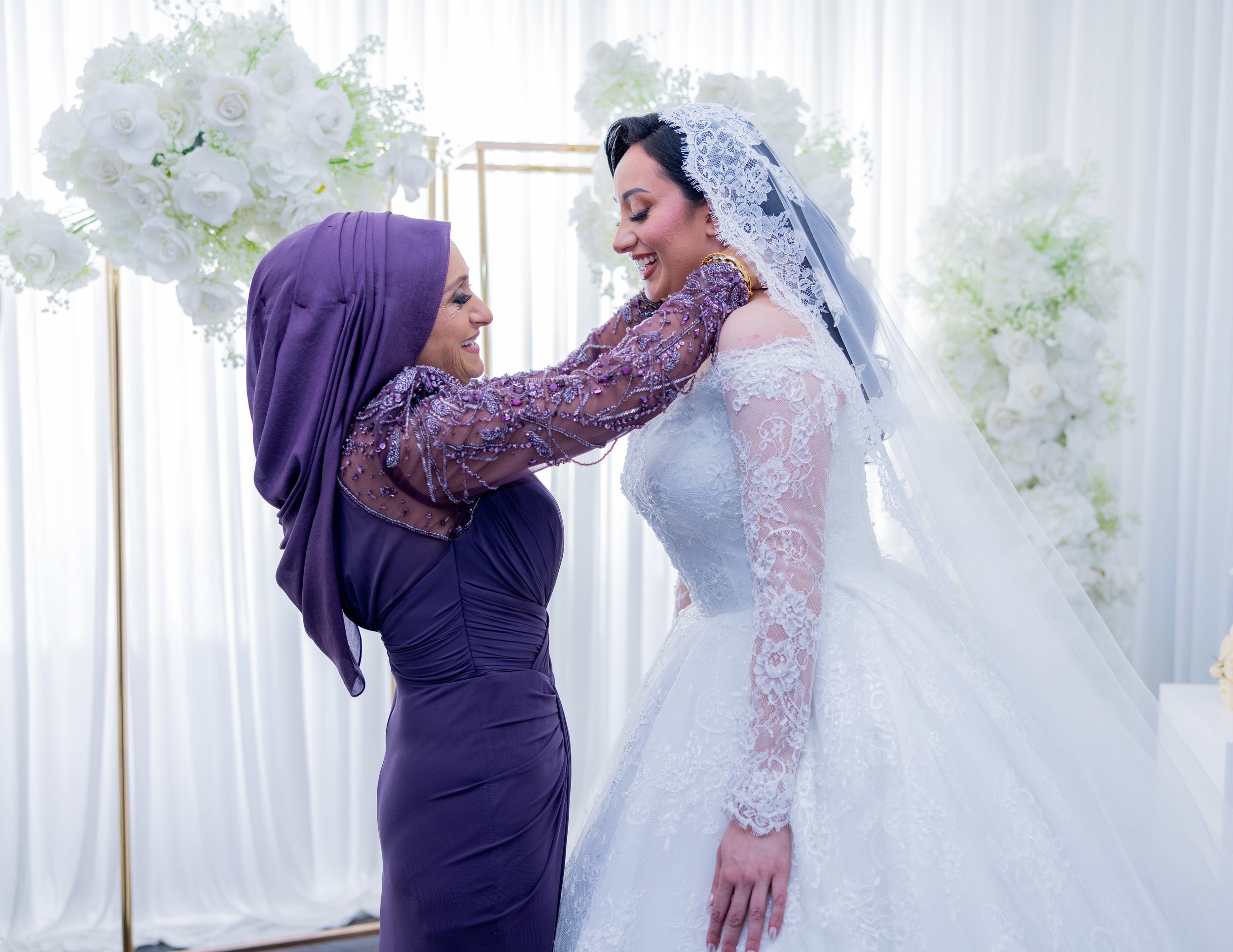 A bride in a white wedding dress and veil smiling as an older woman in a purple dress and headscarf adjusts her jewelry during a wedding ceremony, with white floral arrangements in the background.