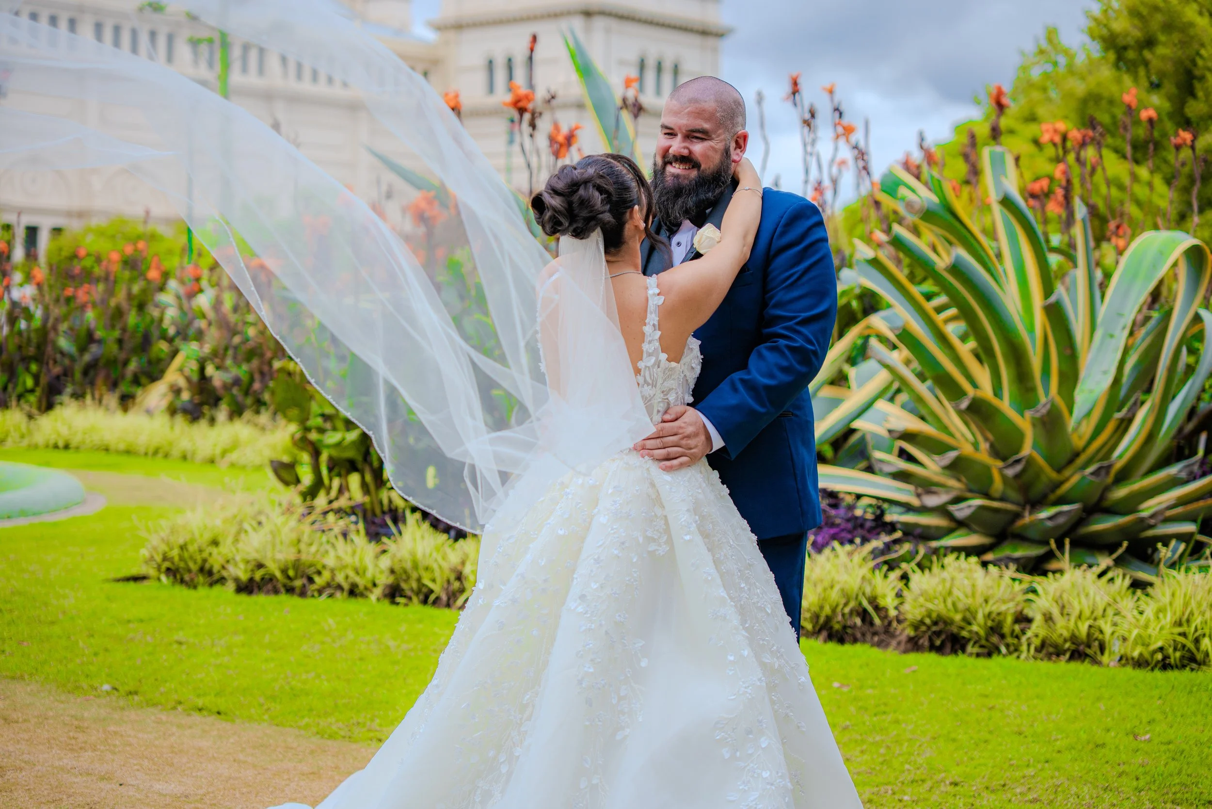 Bride and groom dancing outdoors on their wedding day, surrounded by green plants and large succulent, with a historic building in the background.