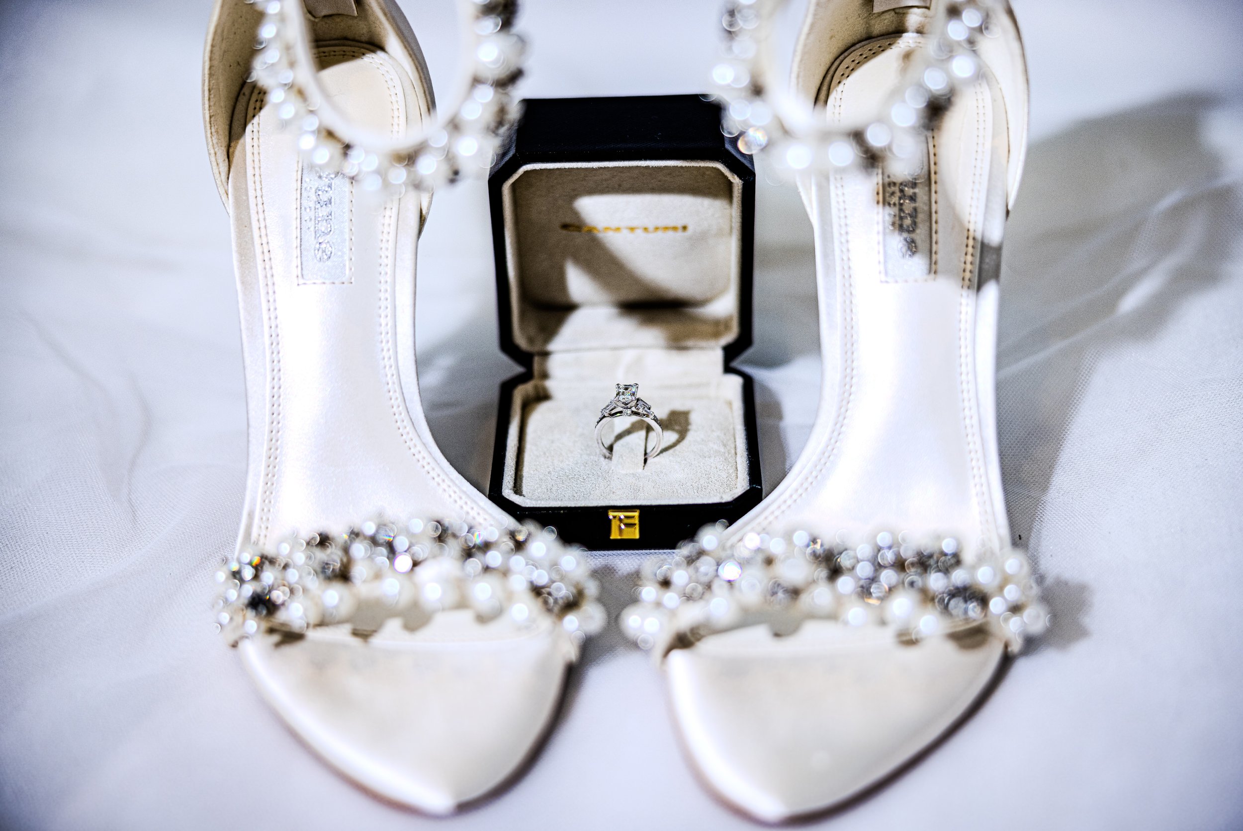 A diamond engagement ring in a jewelry box, flanked by a pair of white high-heeled shoes decorated with pearls and rhinestones.