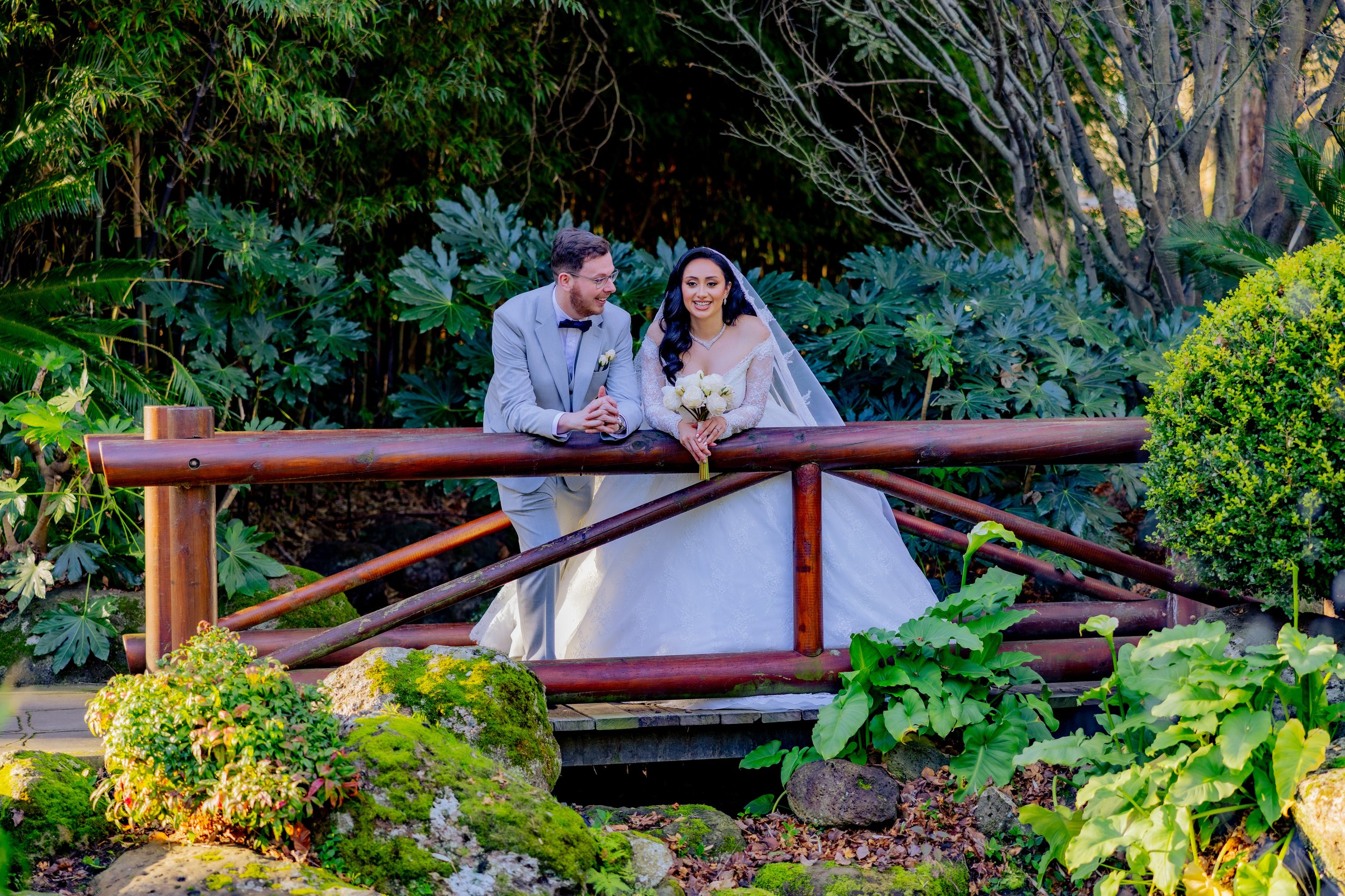 A bride and groom on a wooden bridge in a lush garden, smiling and looking at each other. The bride is holding a bouquet of white roses, and the groom is dressed in a gray suit with a bow tie.